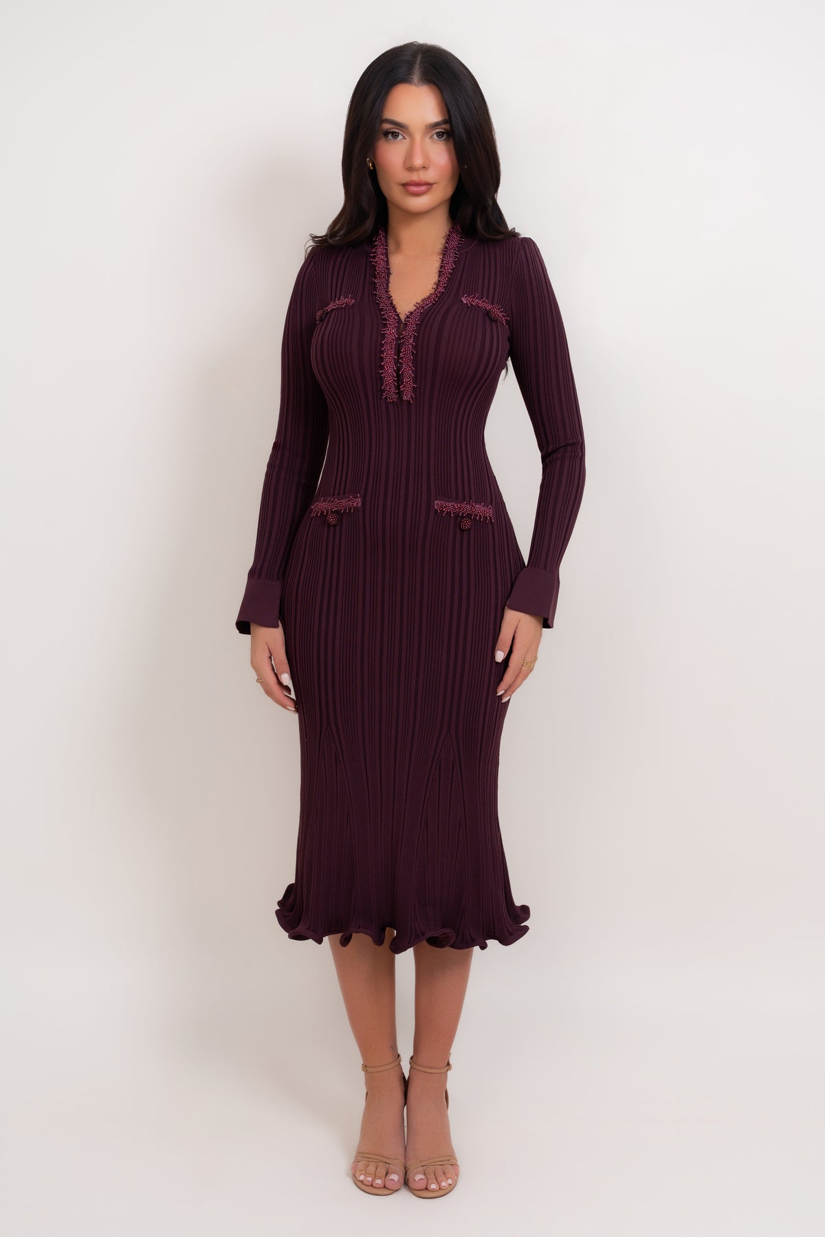 A woman modeling a long-sleeve, dark plum purple ribbed knit midi dress with a ruffled hem and decorative trim details, standing against a plain light background.