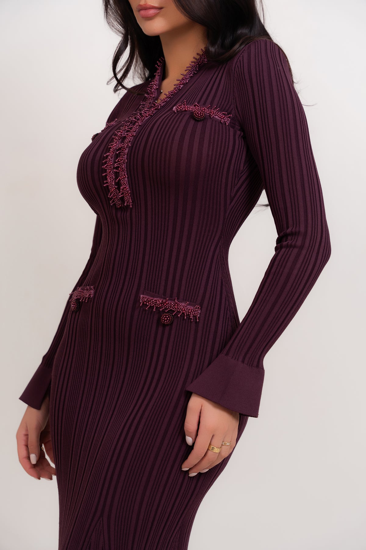 A woman wearing a long-sleeve, bodycon pleated dress in deep plum purple with matching embroidered trim and decorative buttons, shown against a plain light background.