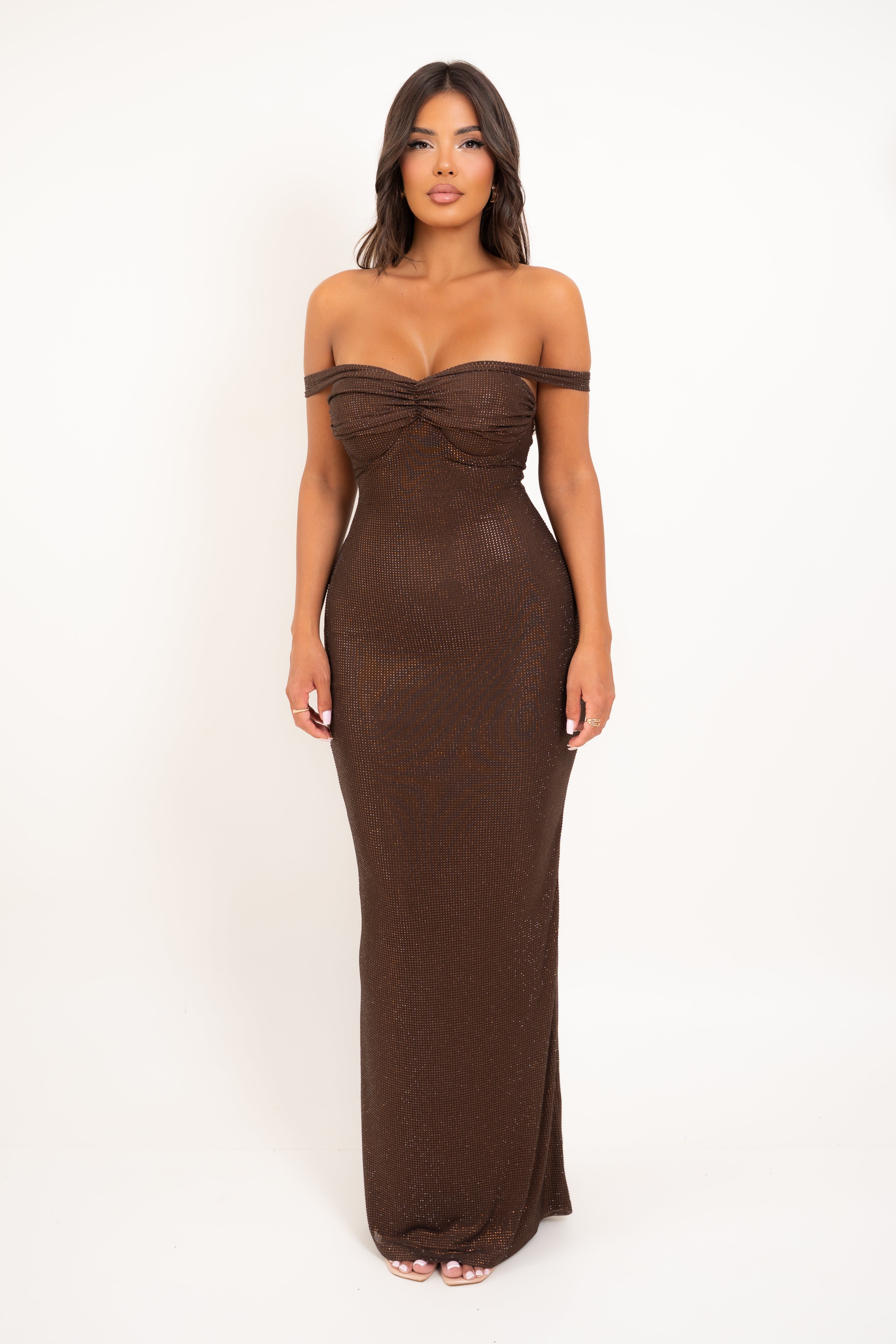A woman modeling a floor-length, off-the-shoulder brown dress with a ruched sweetheart bodice against a plain white studio background.