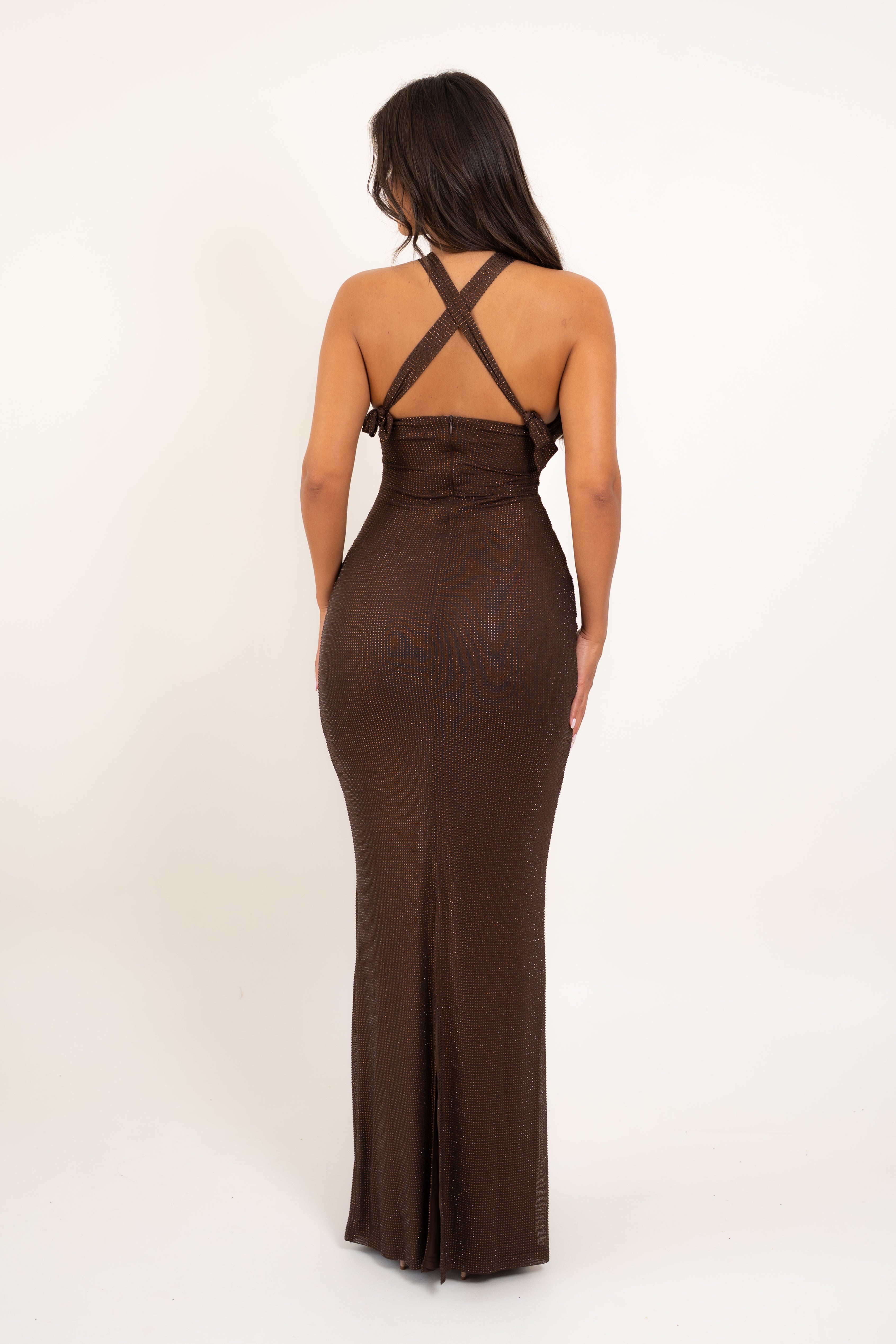A woman viewed from the back wearing a fitted, floor-length brown dress with crisscross shoulder straps and a high waist, photographed against a plain light background.