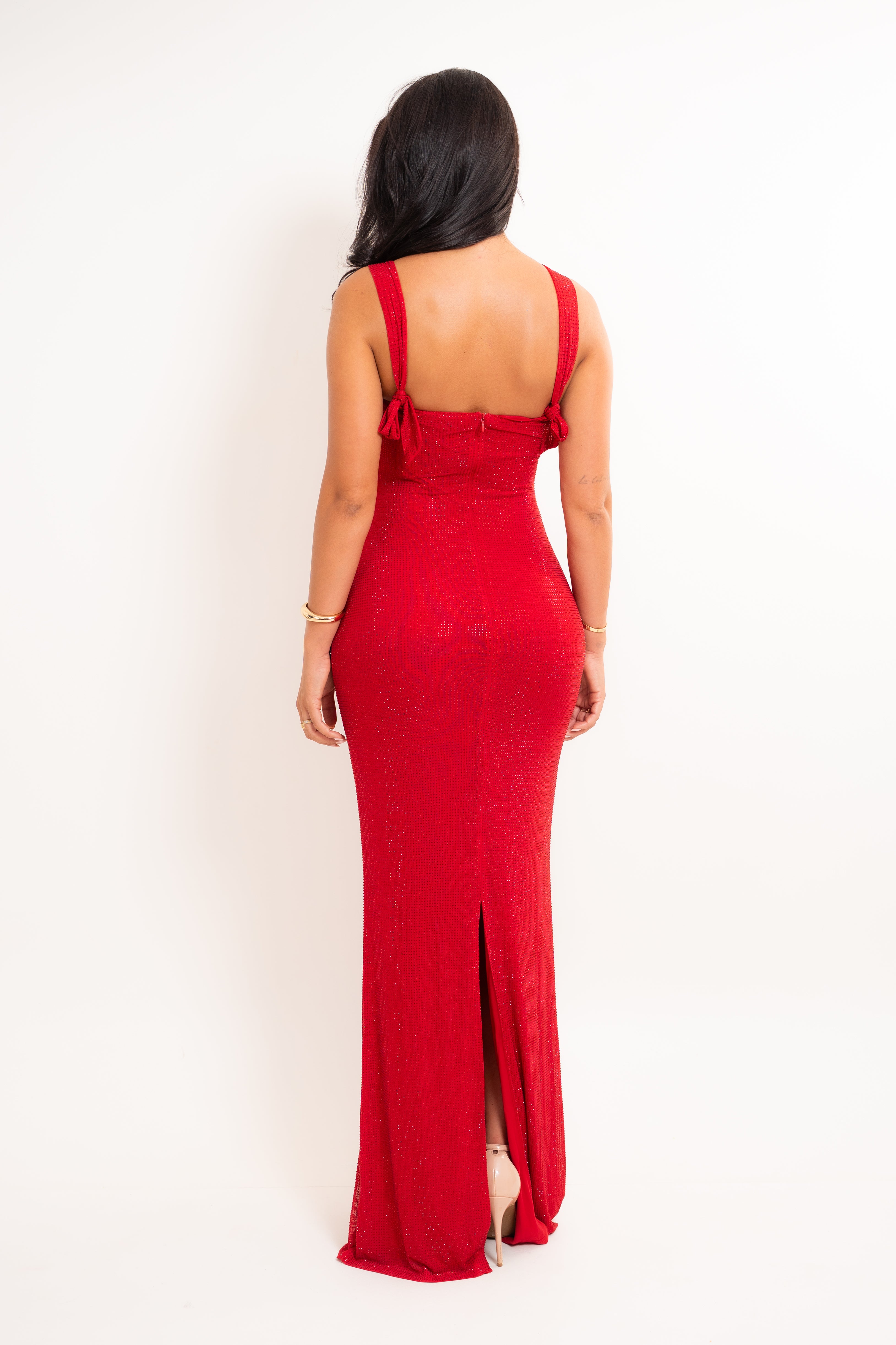 A woman viewed from the back wearing a floor-length red dress with thin shoulder straps, a fitted bodice, and a front slit, standing against a plain light background.