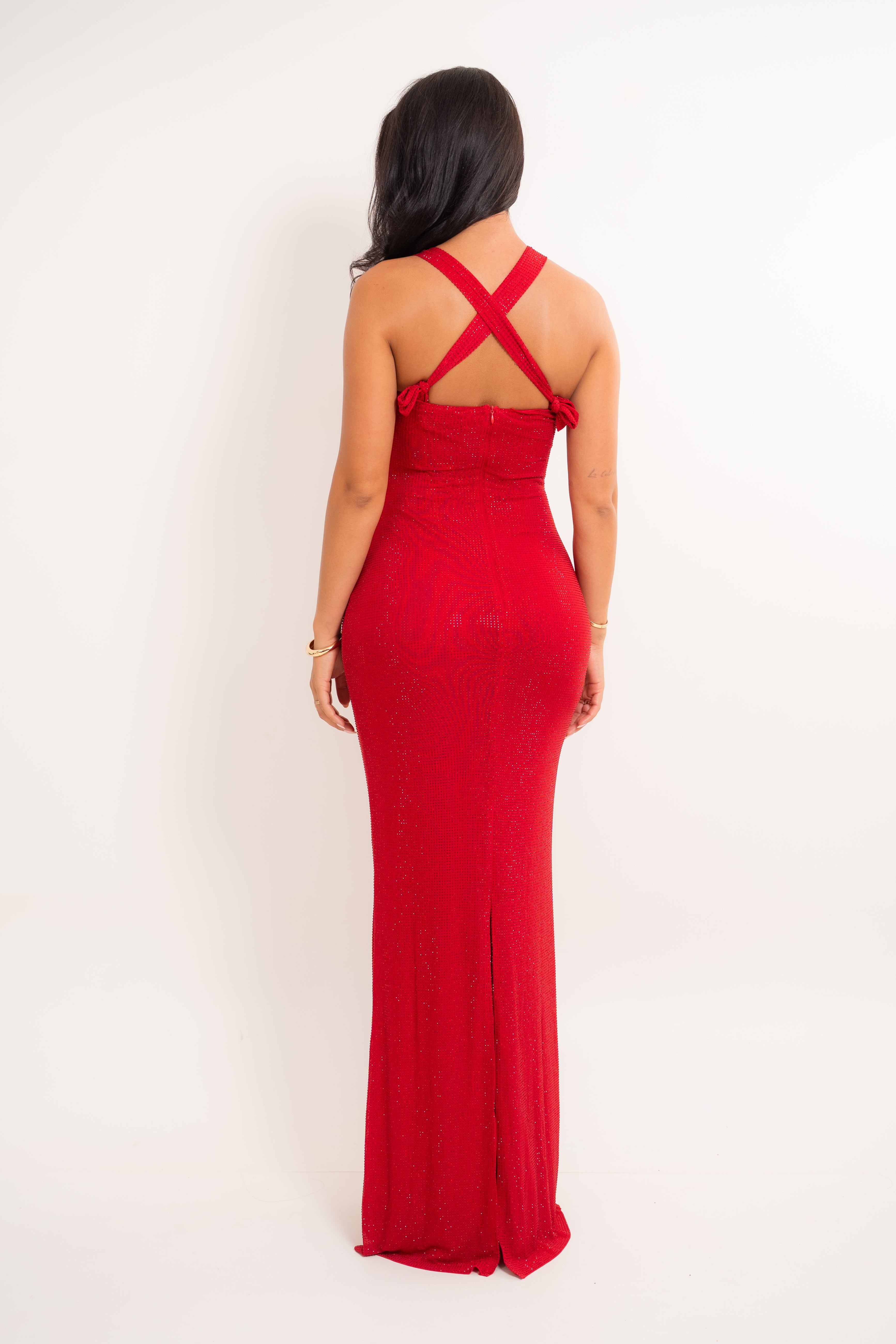 A woman wearing a floor-length red dress, shown from the back against a plain white studio background. The dress has crisscross back straps and a fitted silhouette.