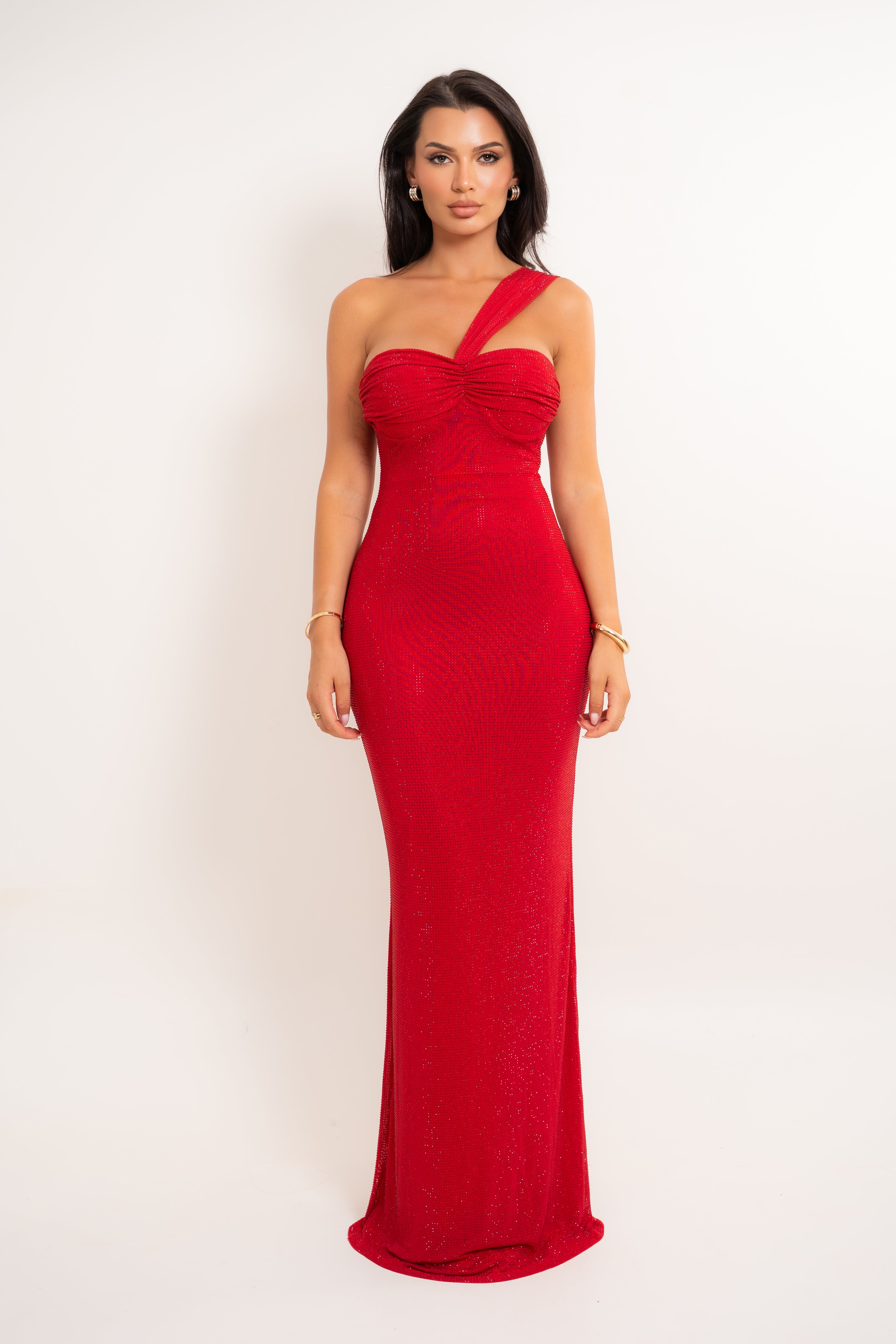 A woman modeling a floor-length, one-shoulder red gown with a ruched sweetheart bodice and fitted silhouette, photographed against a plain light background.