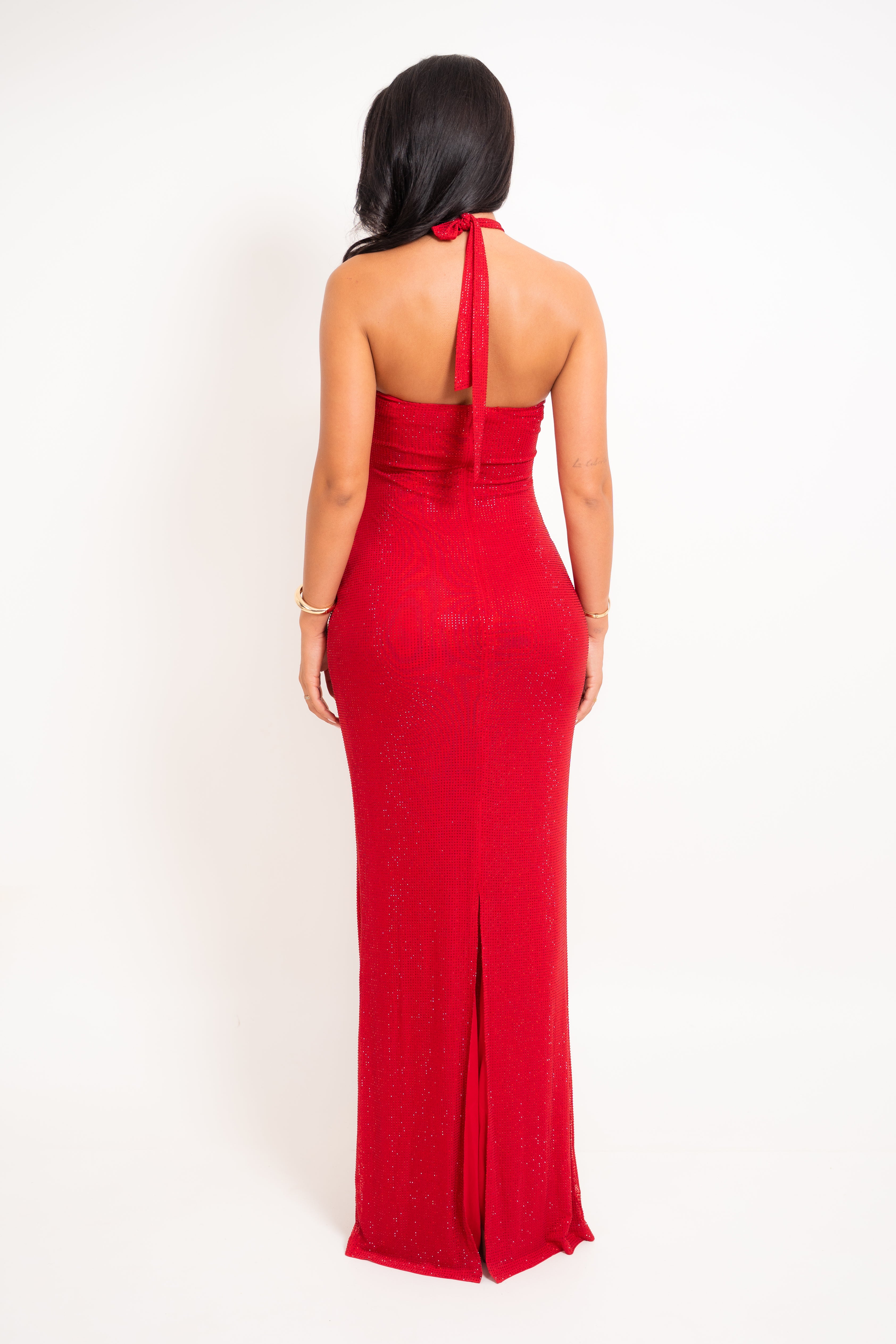 A woman viewed from the back wearing a sleeveless red halter maxi dress with a fitted silhouette and a front slit, standing against a plain white studio background.