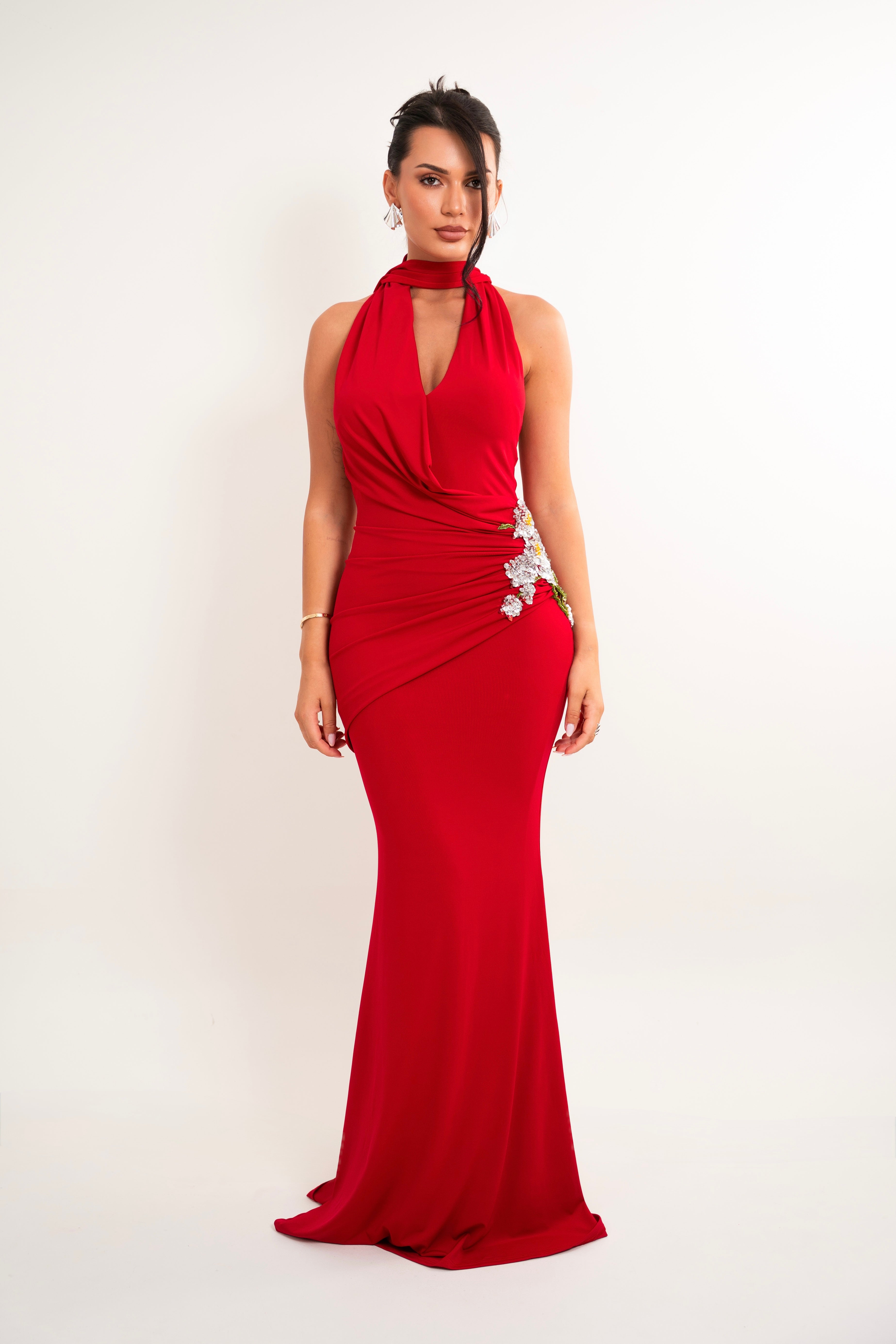 A woman modeling a floor-length red halter gown with a draped neckline, fitted silhouette, and floral embellishment on the hip, photographed against a plain light background.