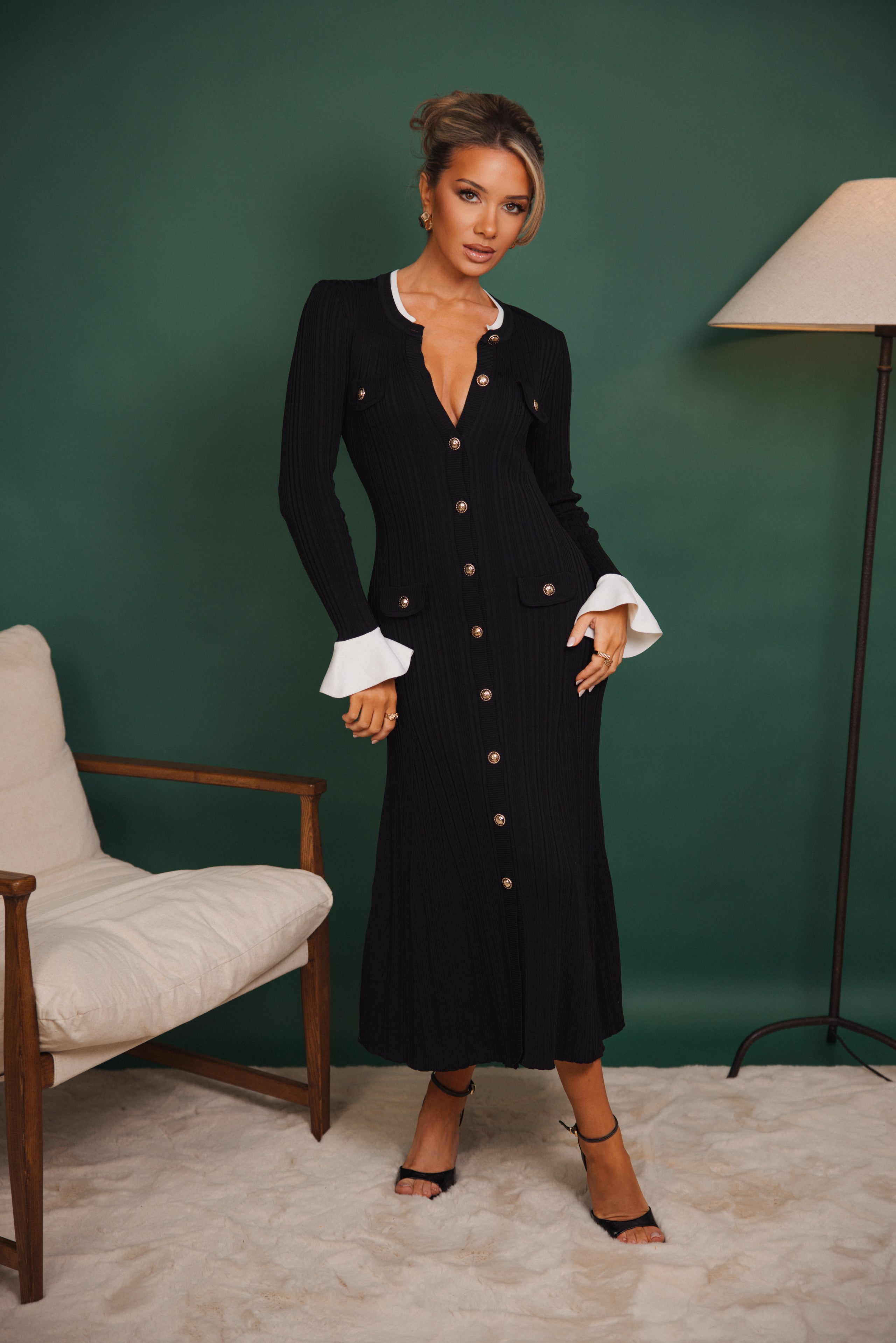 Woman in a black dress with white cuffs standing against a green wall.