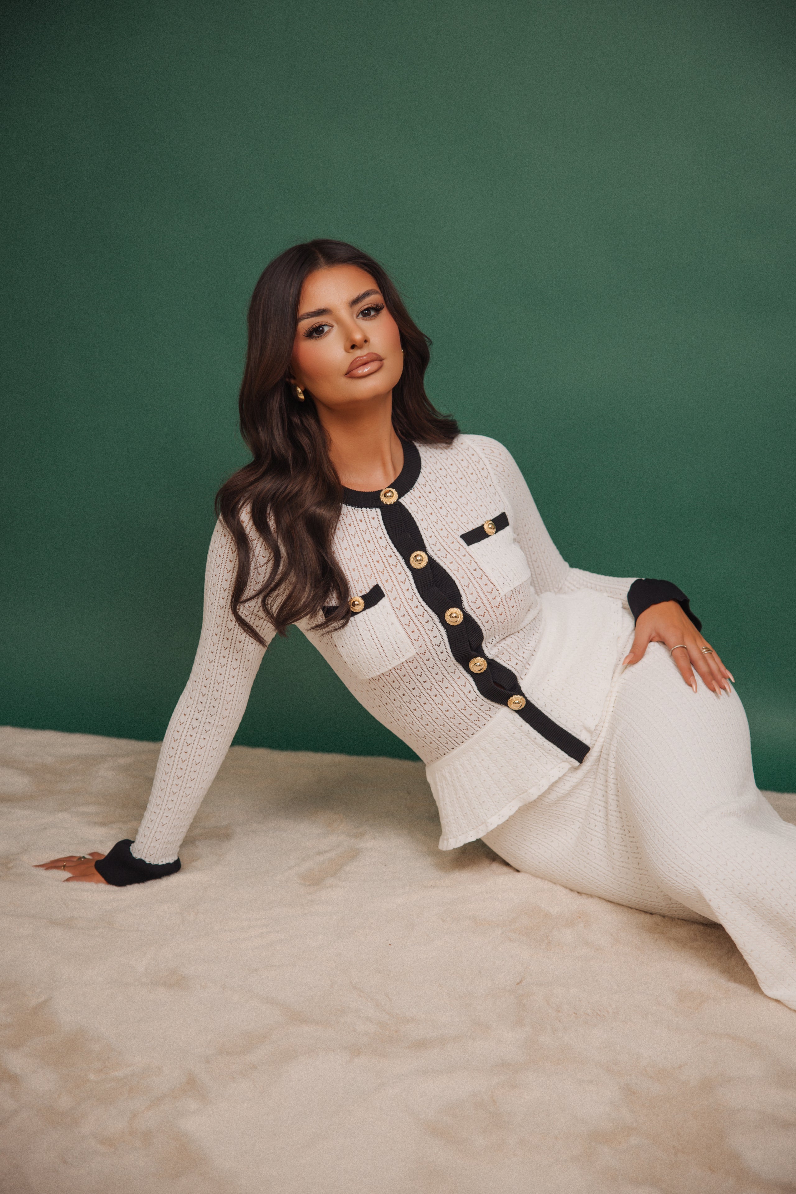 Woman in a white outfit with black accents sitting on a beige surface against a green background