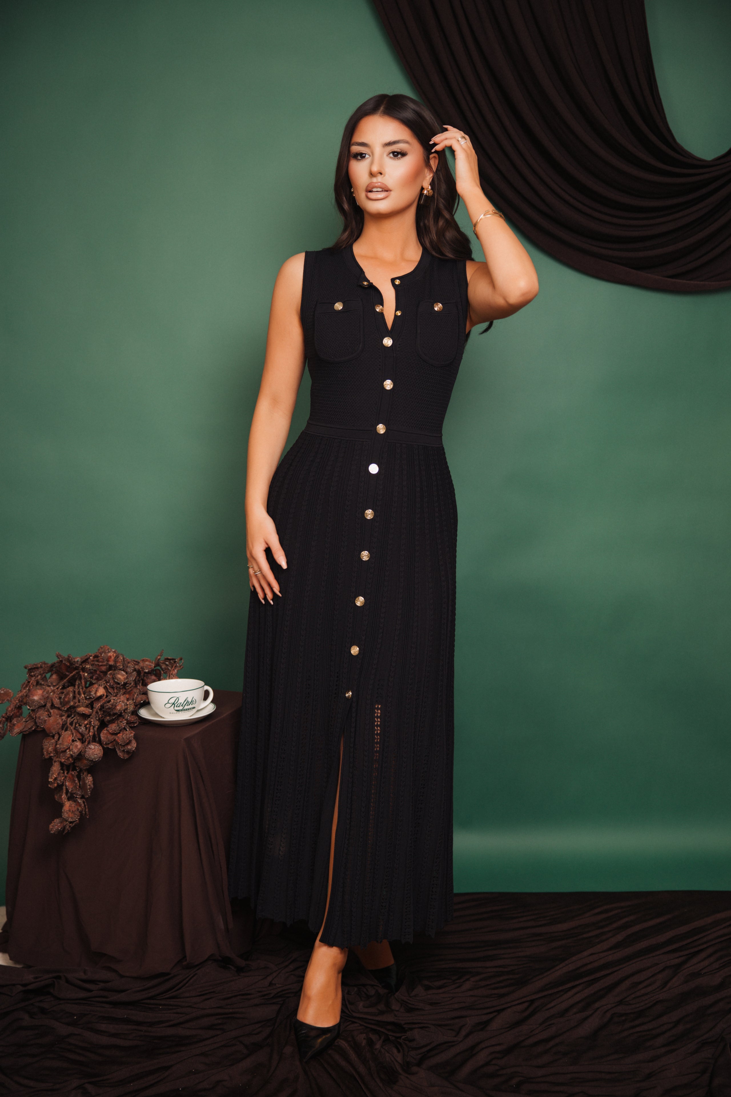 Woman in a black dress standing against a green wall with a table and decorative items.