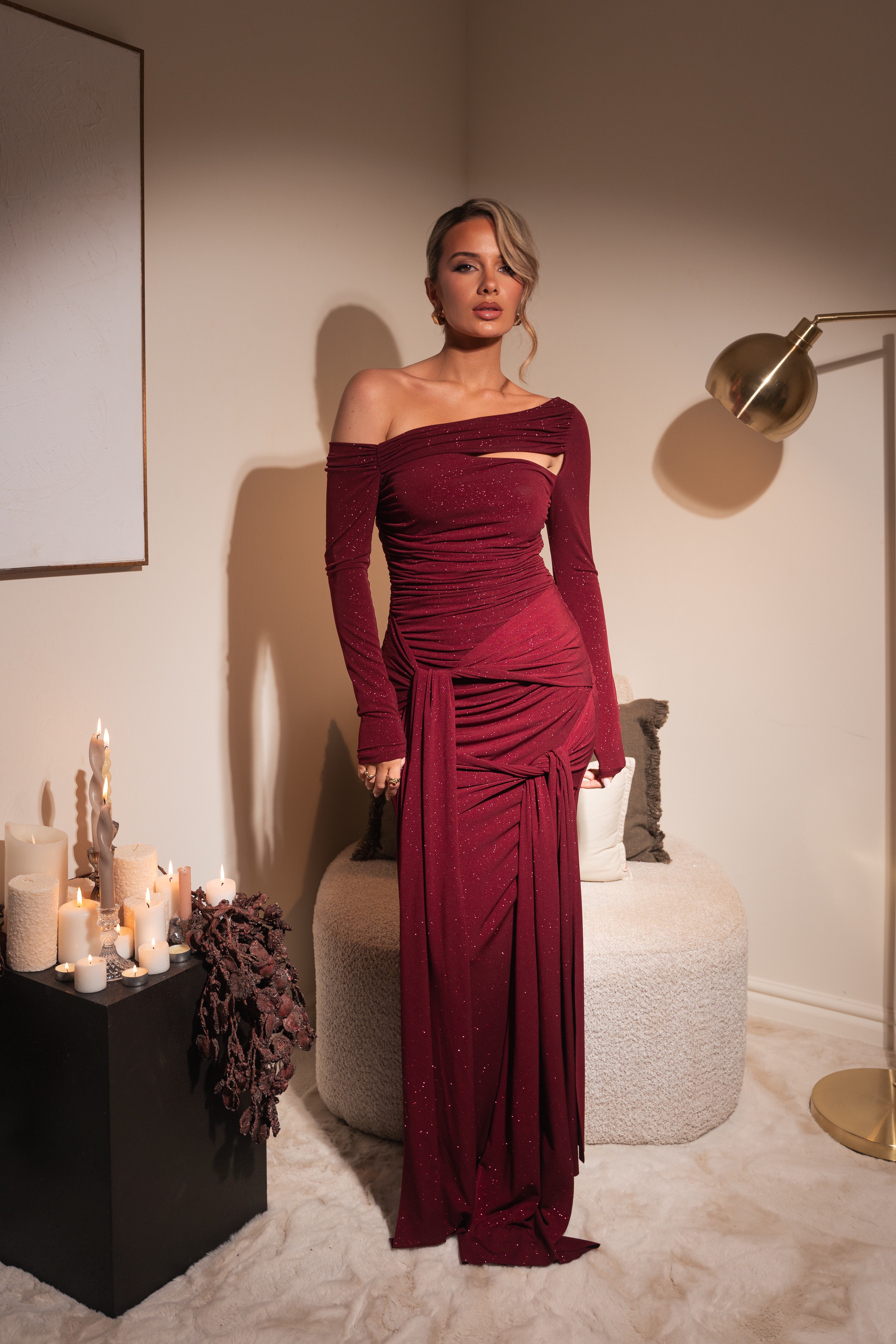 Woman in a burgundy off-shoulder dress standing in an elegant room with candles and decor.