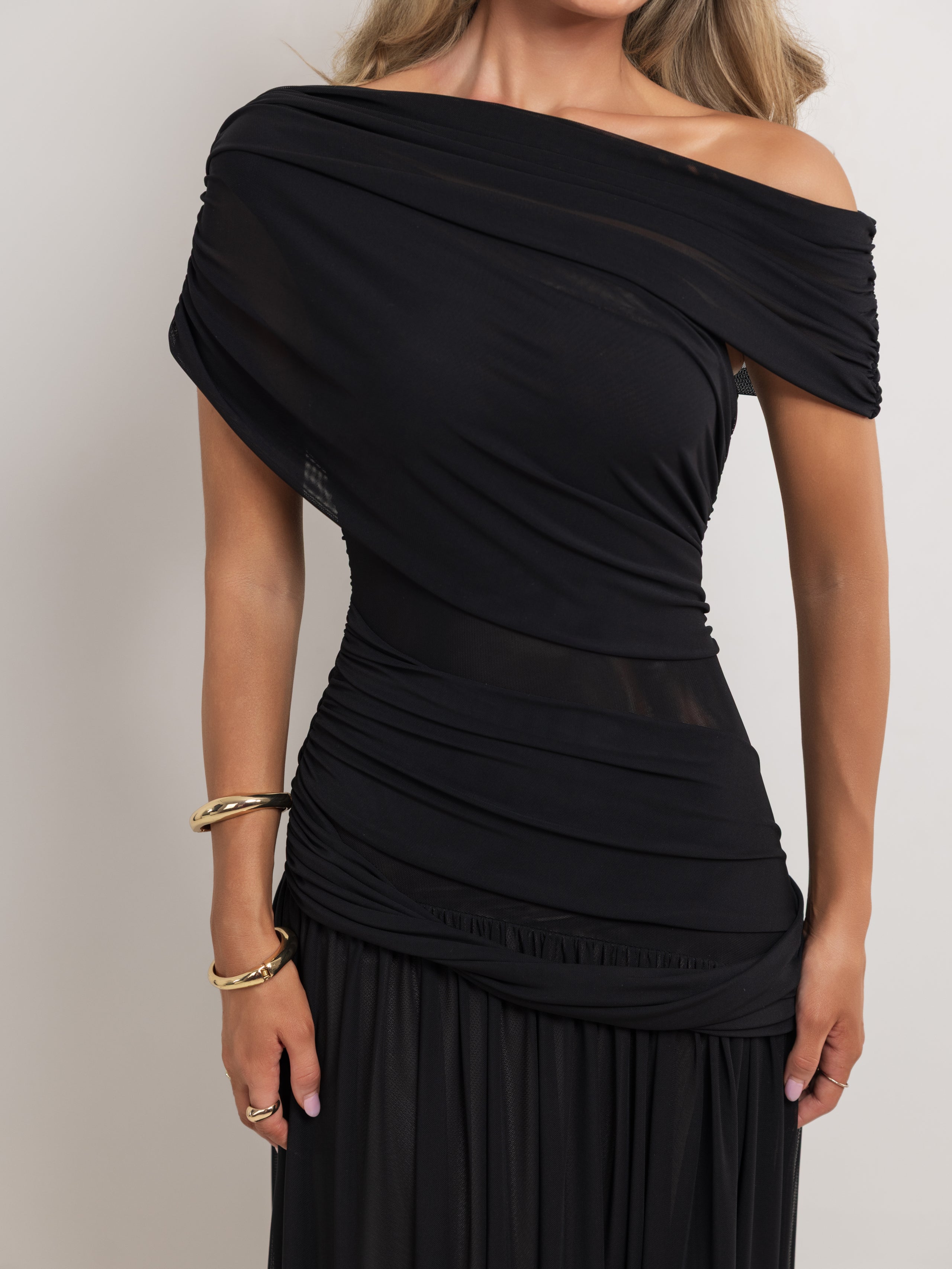A woman wearing a black off-the-shoulder draped dress with ruching and a pleated skirt, shown against a plain light gray background.