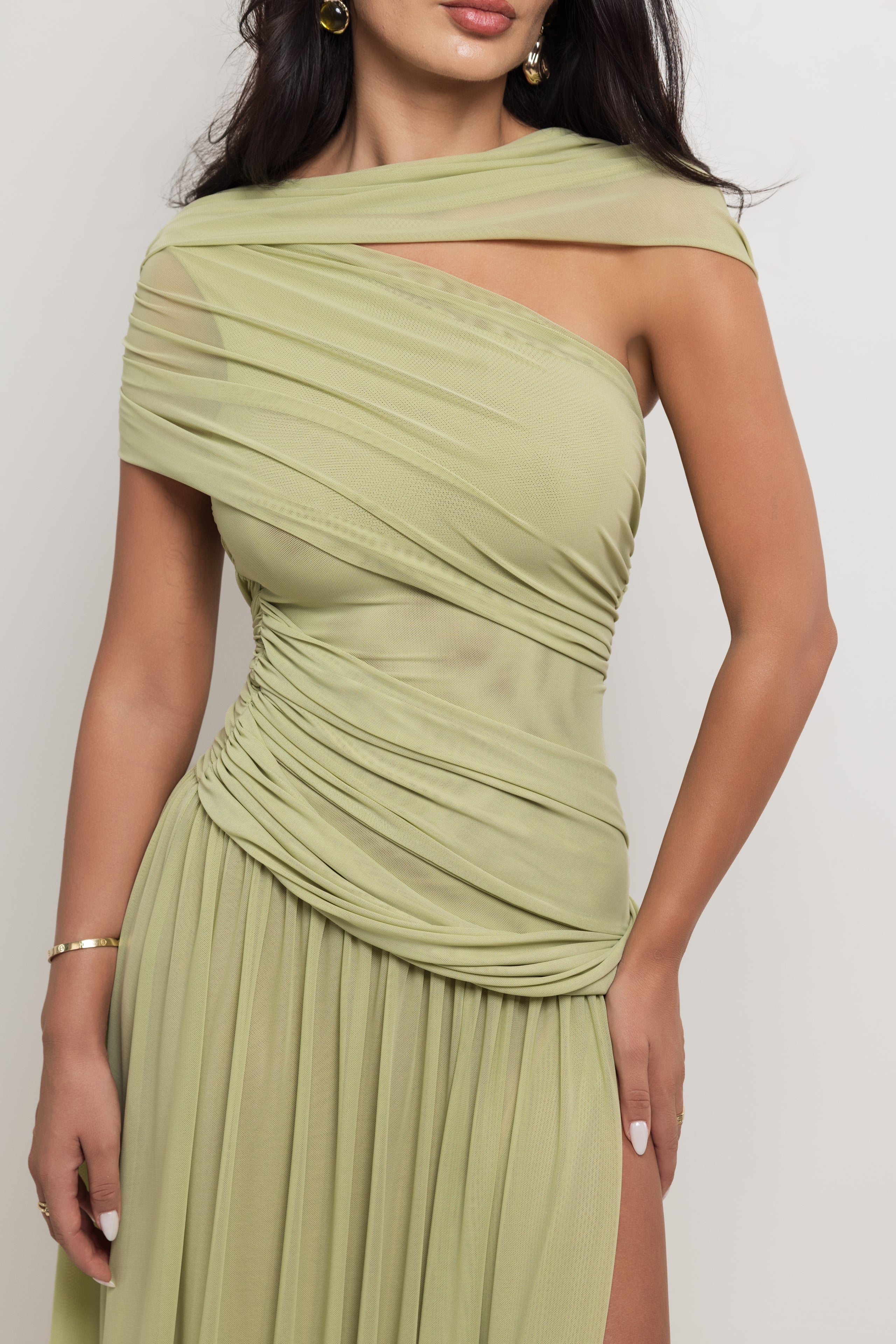 A woman wearing a sage green one-shoulder draped dress with a pleated skirt, shown from the chest down against a light gray studio background.