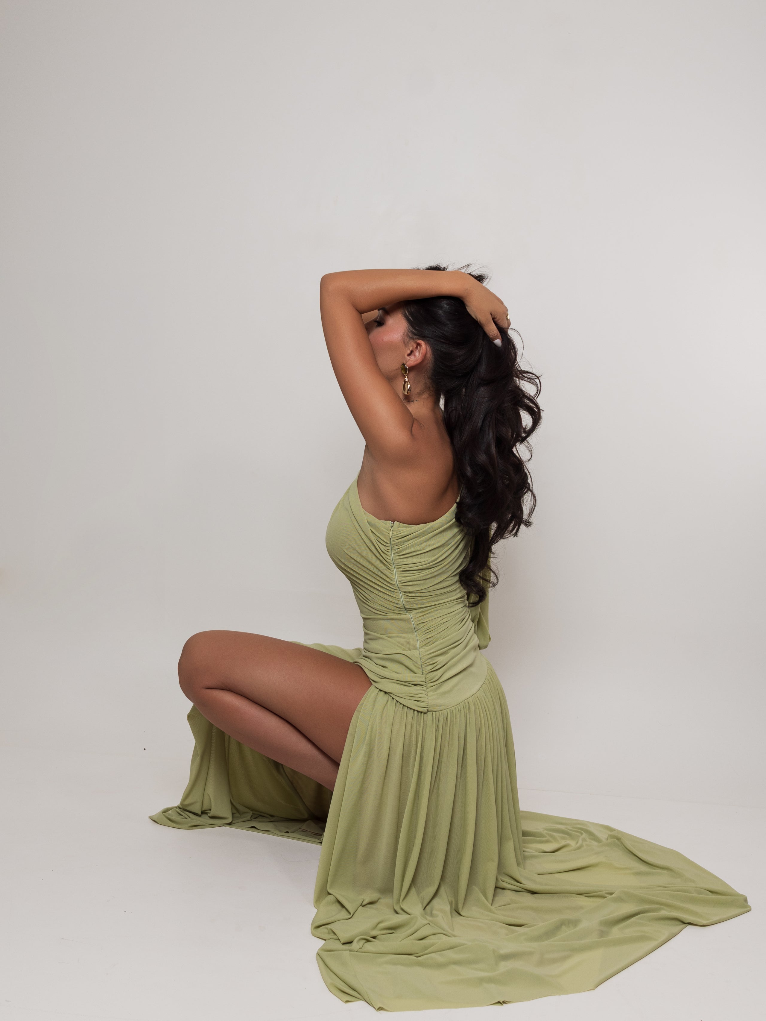 A woman kneels on a white studio floor wearing a sage green ruched maxi dress with a high side slit and a long flowing train.