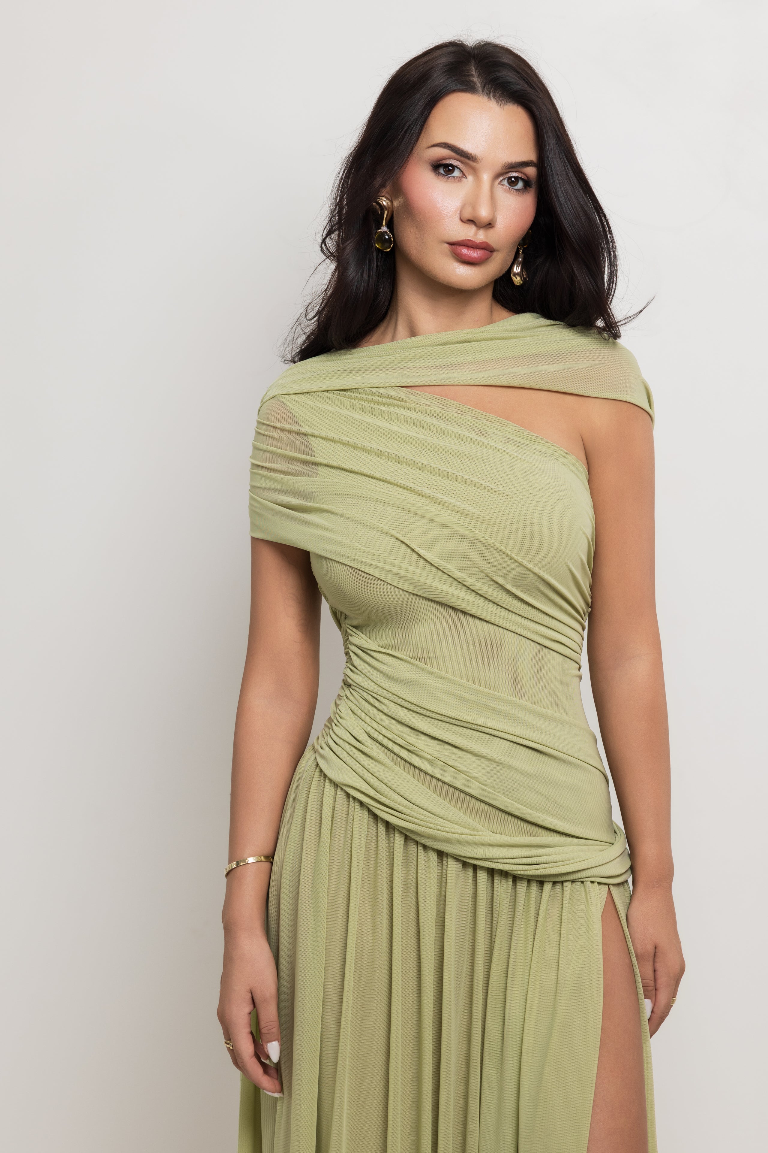 A woman modeling a draped one-shoulder midi dress in a muted sage green color, featuring sheer mesh panels and a high side slit, set against a plain light gray background.