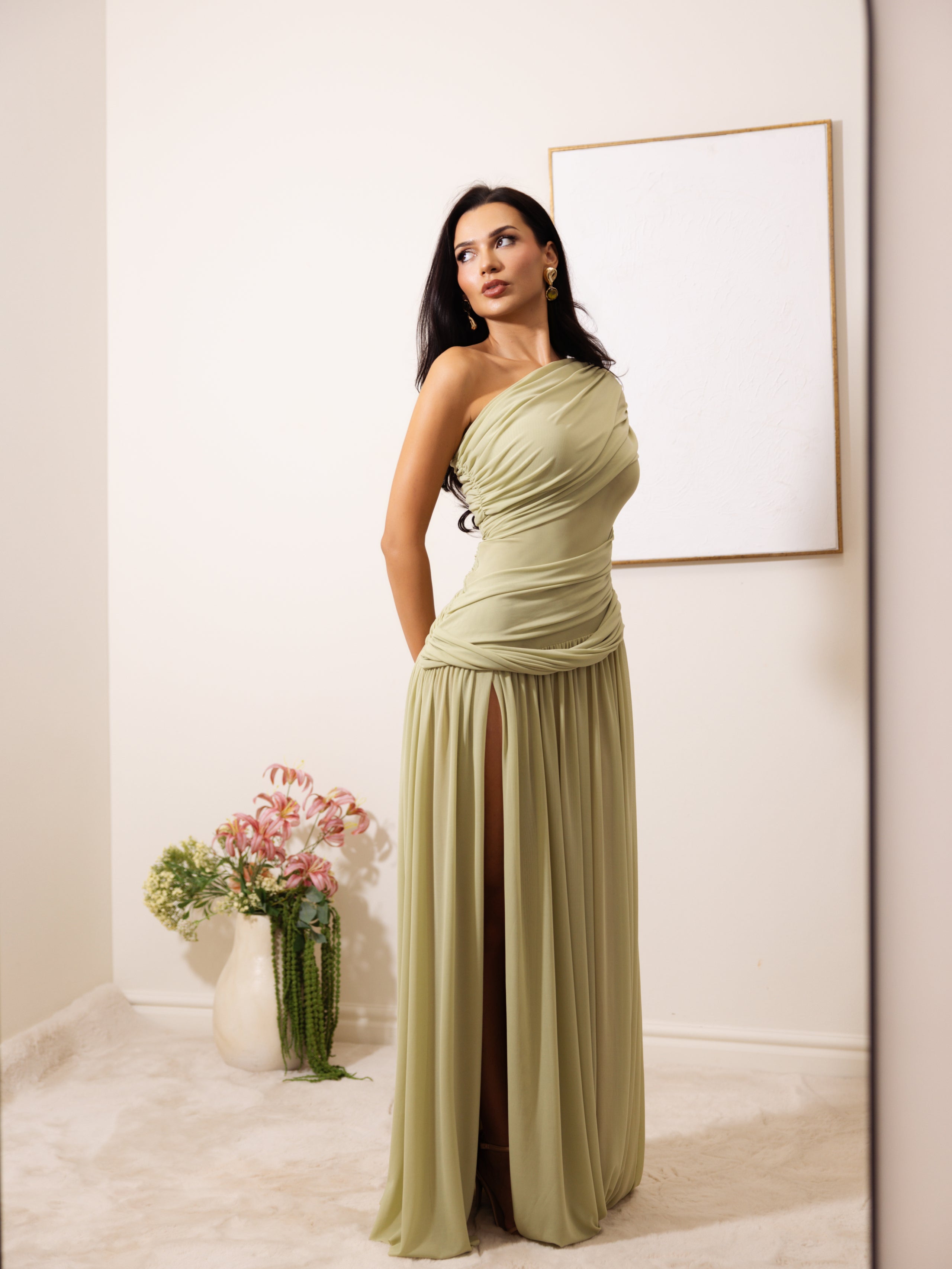 A woman models a one-shoulder sage green maxi dress with a ruched bodice and a high thigh slit, standing in a minimalist room with a blank framed picture on the wall and a vase of flowers on the floor.
