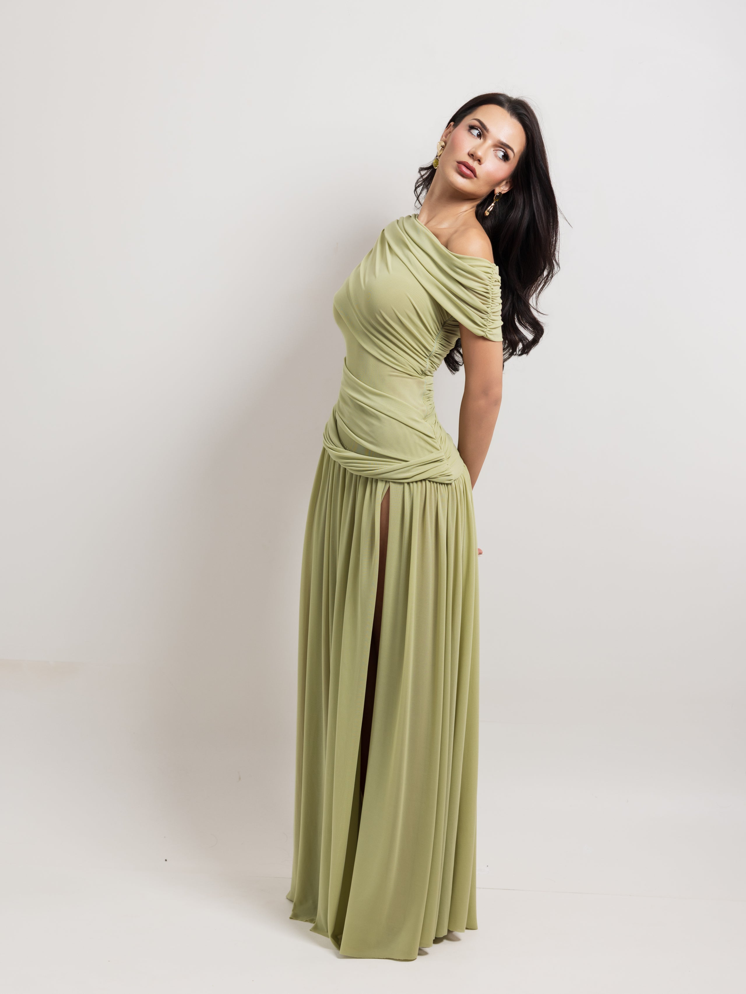 A woman modeling a floor-length olive green off-the-shoulder gown with ruching on the bodice and a high side slit, photographed against a plain light gray studio background.