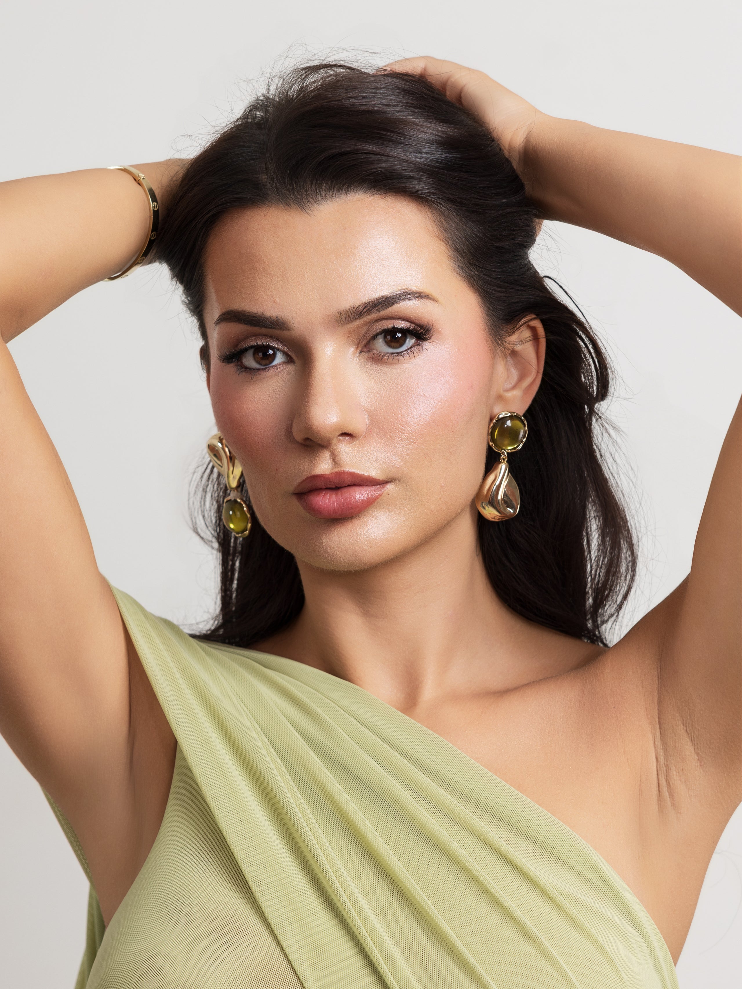 A woman with dark hair pulled back wears large gold and olive green teardrop earrings, with her hands in her hair. She is wearing a light sage green draped top against a plain light gray background.