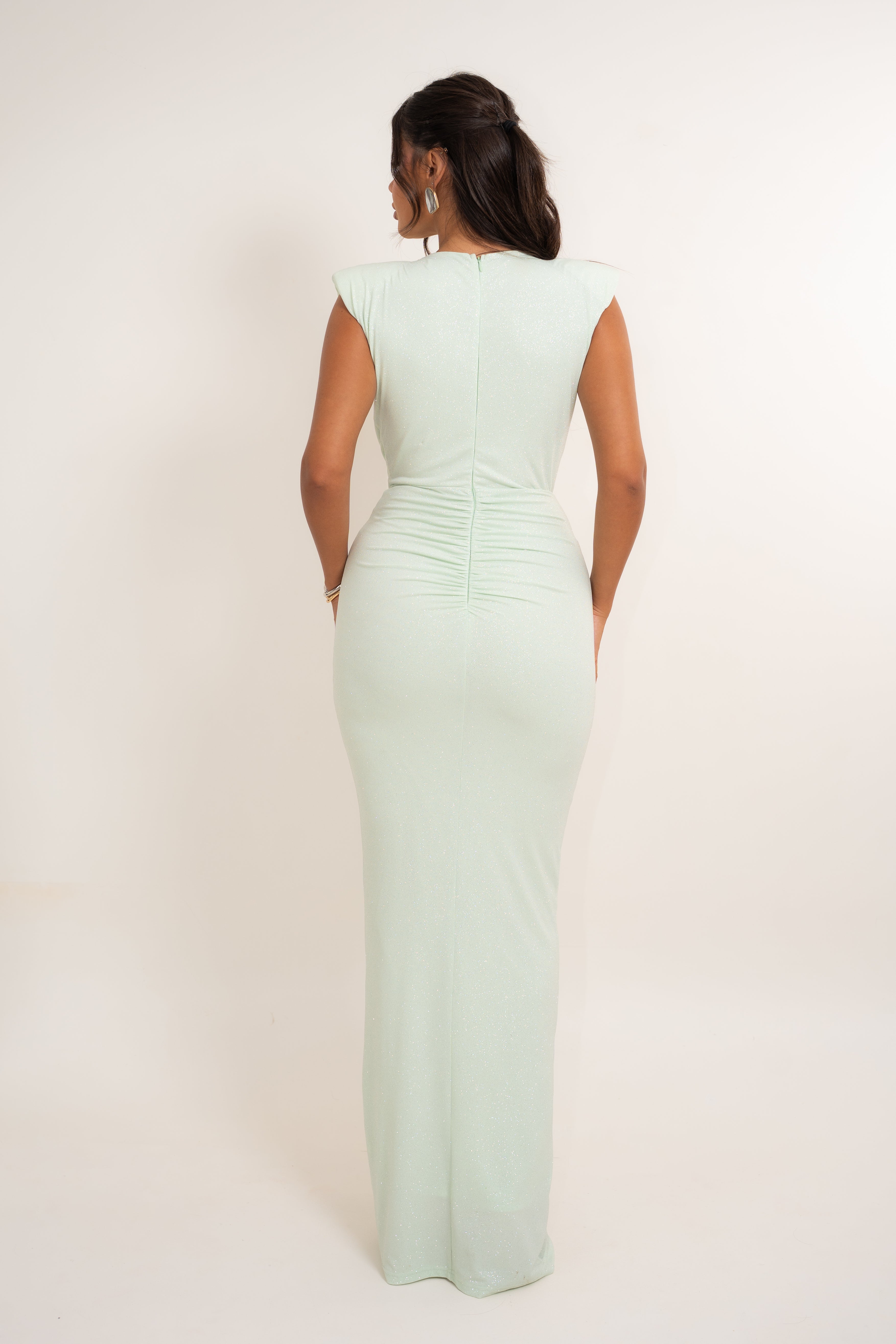 A woman viewed from the back wearing a pale mint green bodycon maxi dress with cap sleeves and a ruched waist, photographed against a plain light beige studio background.