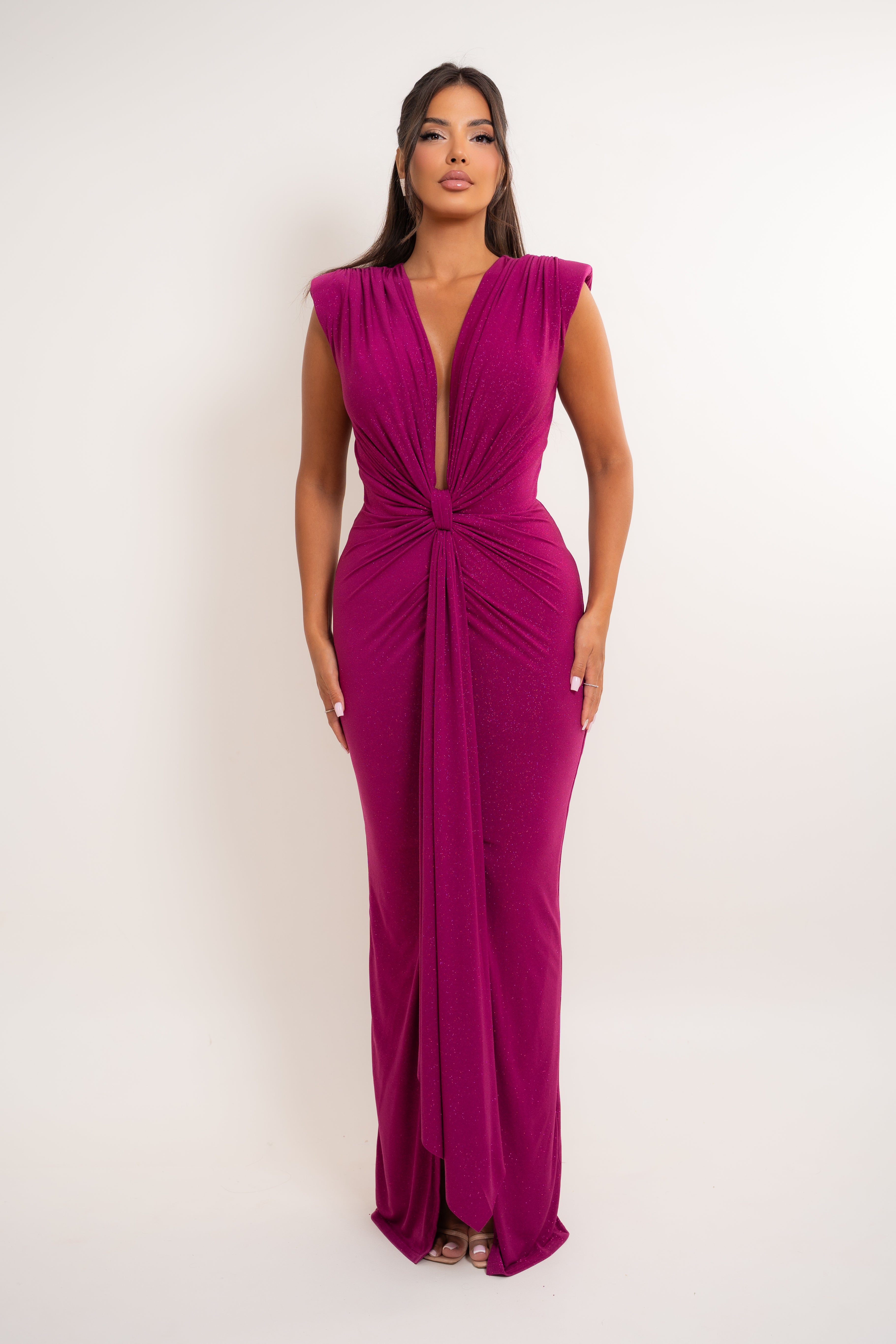 A woman modeling a sleeveless magenta maxi dress with a plunging V-neckline, twisted waist detail, and side pockets, set against a plain light gray studio background.