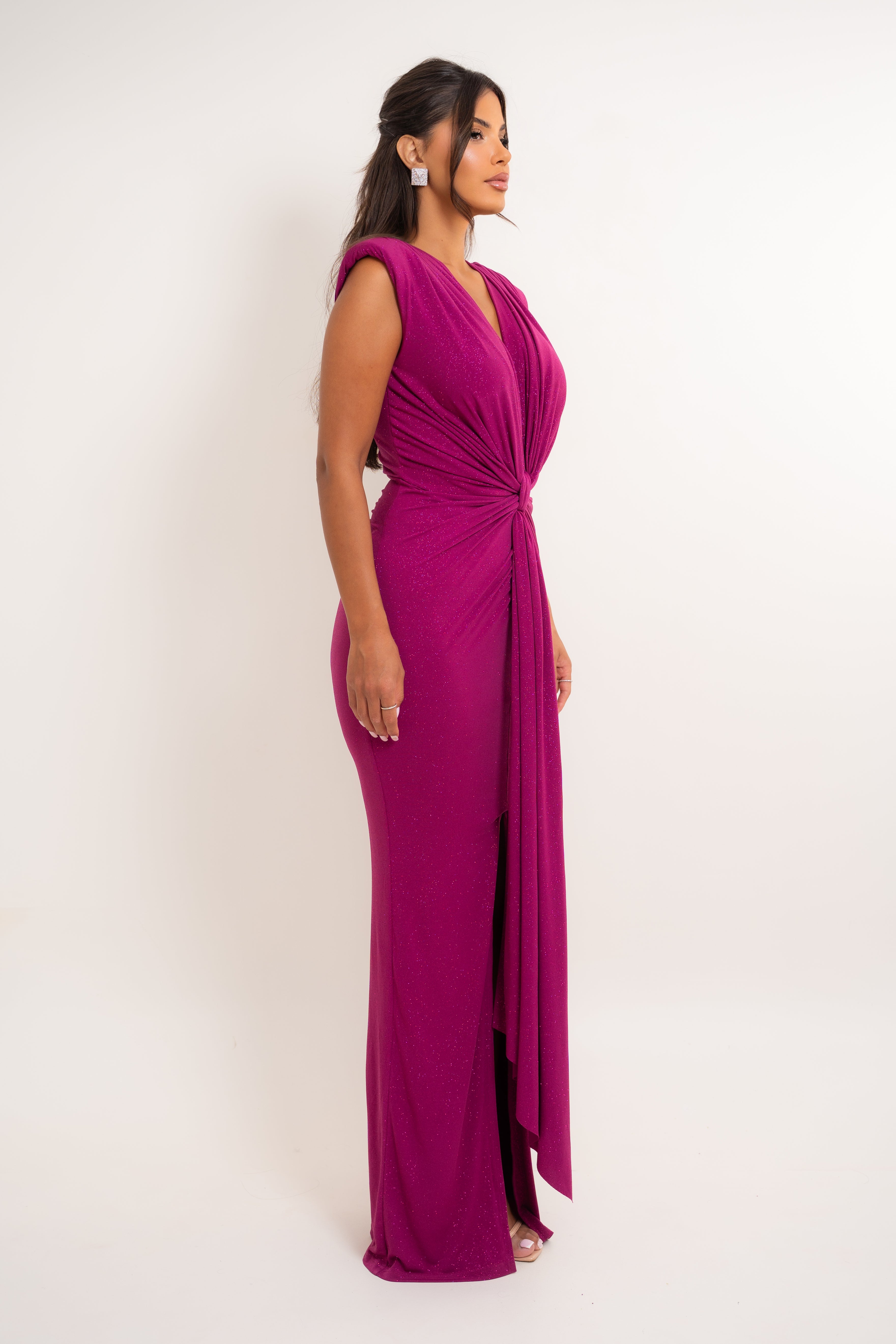 A woman modeling a sleeveless, deep V-neck floor-length gown in a rich magenta-purple color. The dress features a twisted knot detail at the waist and a high-low draped hemline with a front slit, shown against a plain white studio background.