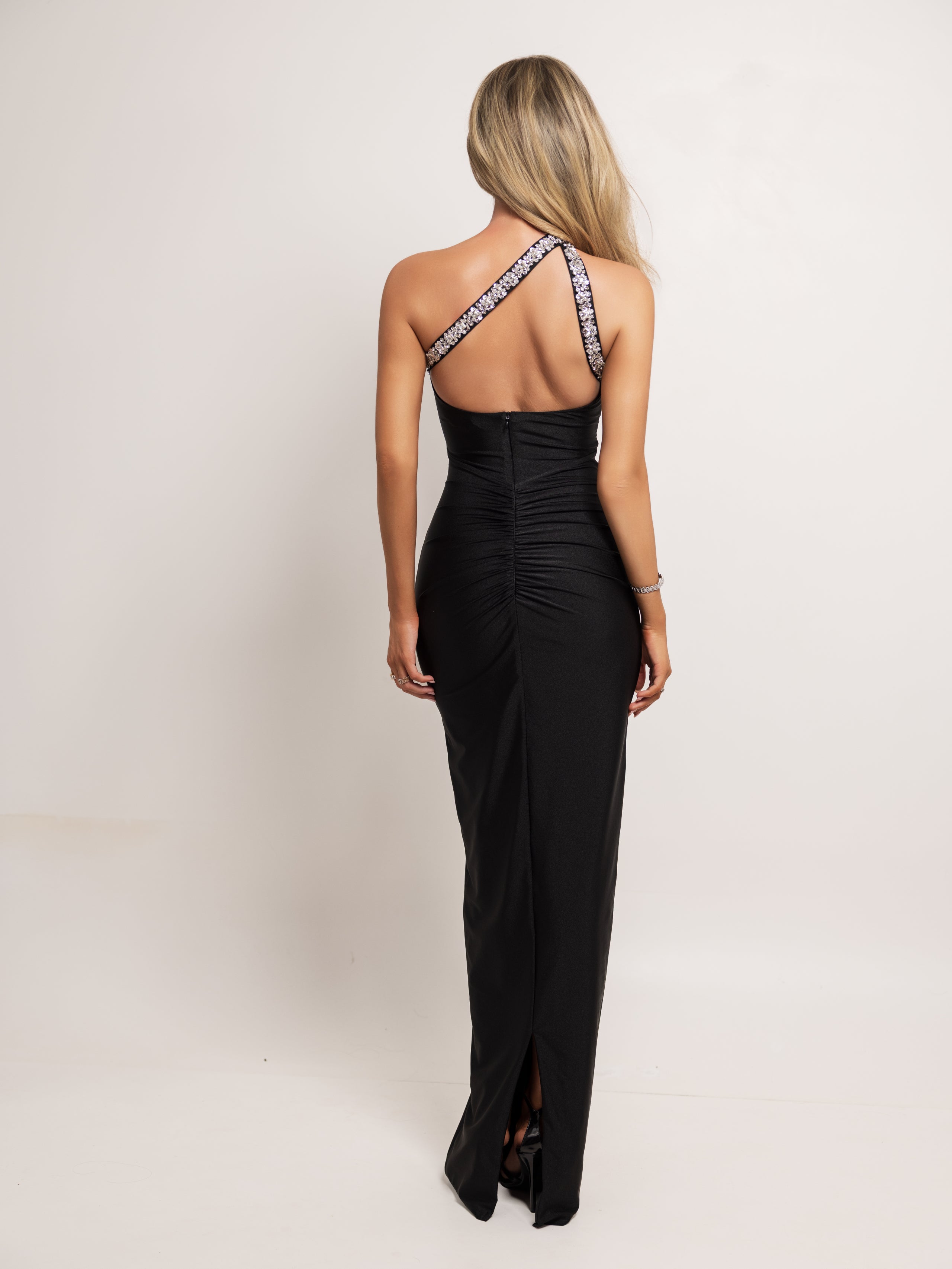 A woman viewed from the back wearing a black floor-length gown with a ruched bodice and patterned halter-style shoulder straps, photographed against a plain light studio background.