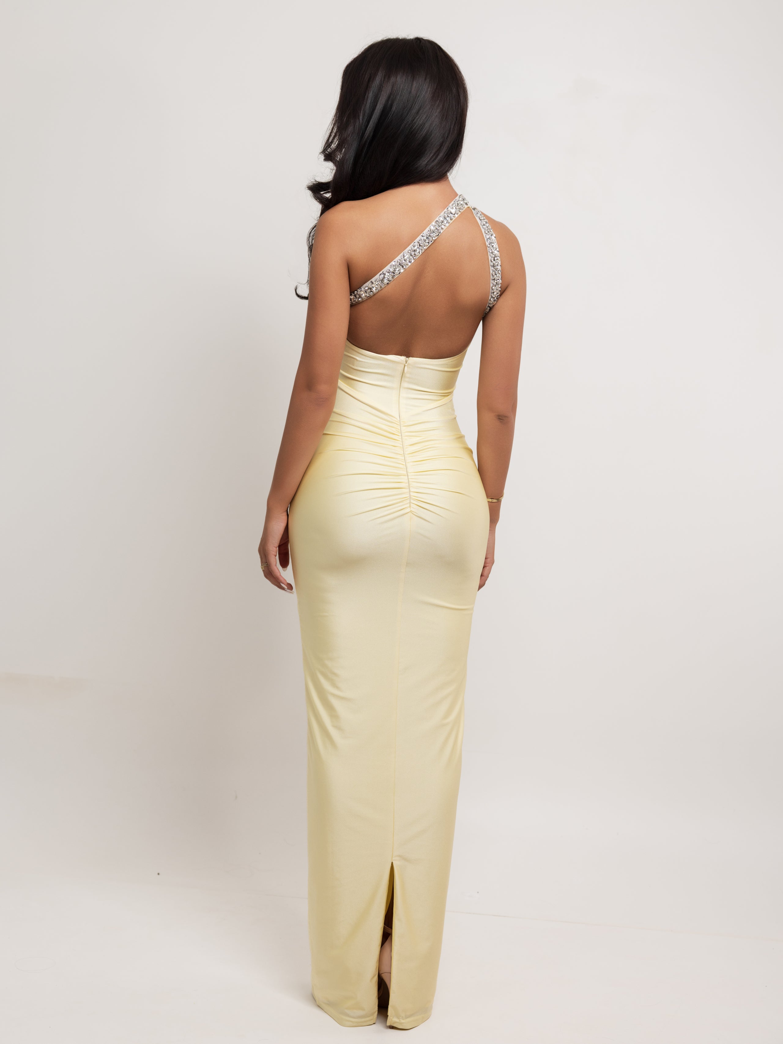A woman shown from the back wearing a pale yellow, form-fitting maxi dress with a low open back, ruched waist, and sparkling silver embellished shoulder straps, set against a plain white studio background.