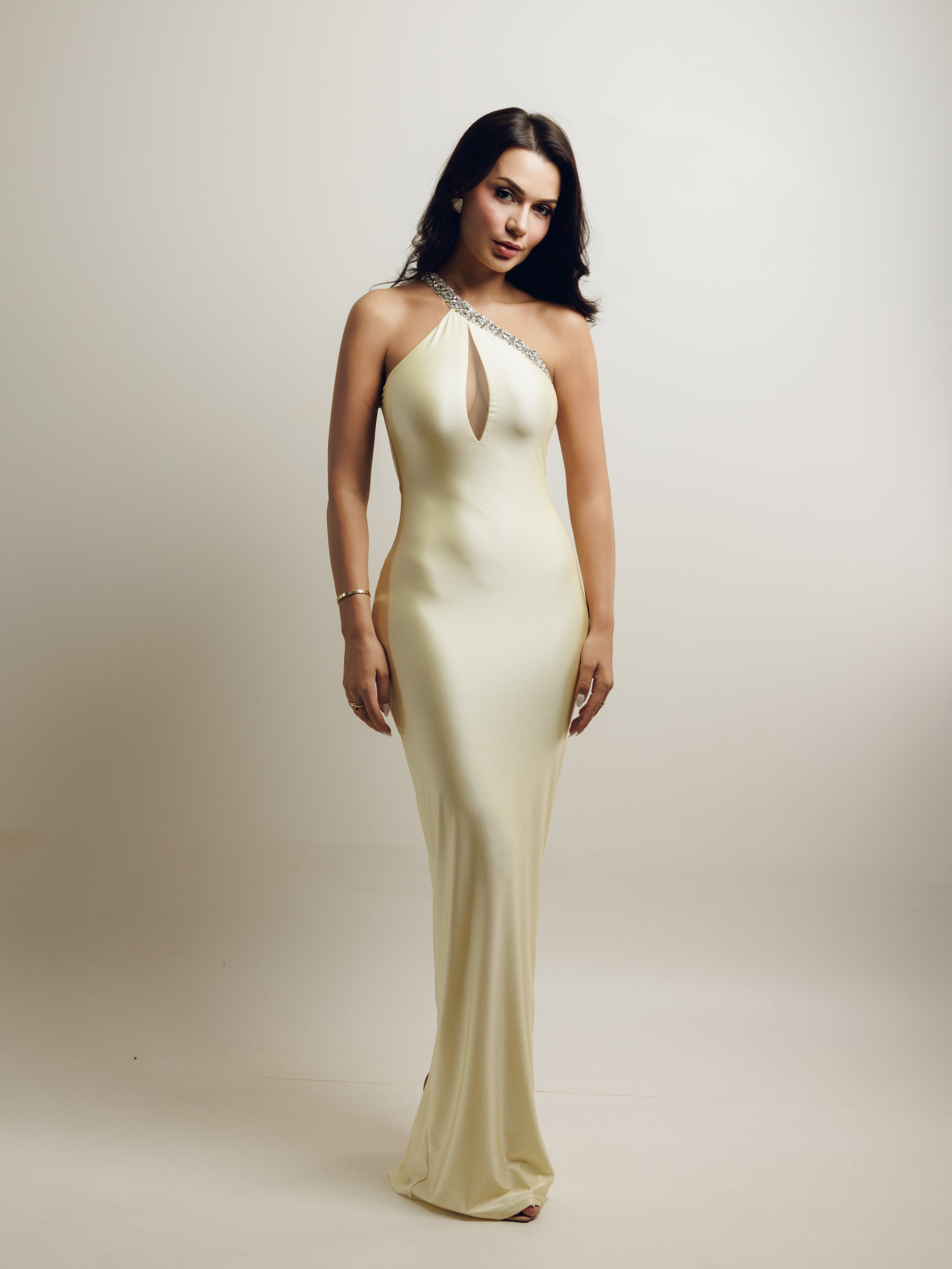 A woman modeling a floor-length, one-shoulder champagne gown with a keyhole cutout and crystal embellishments along the neckline, set against a plain light beige studio background.