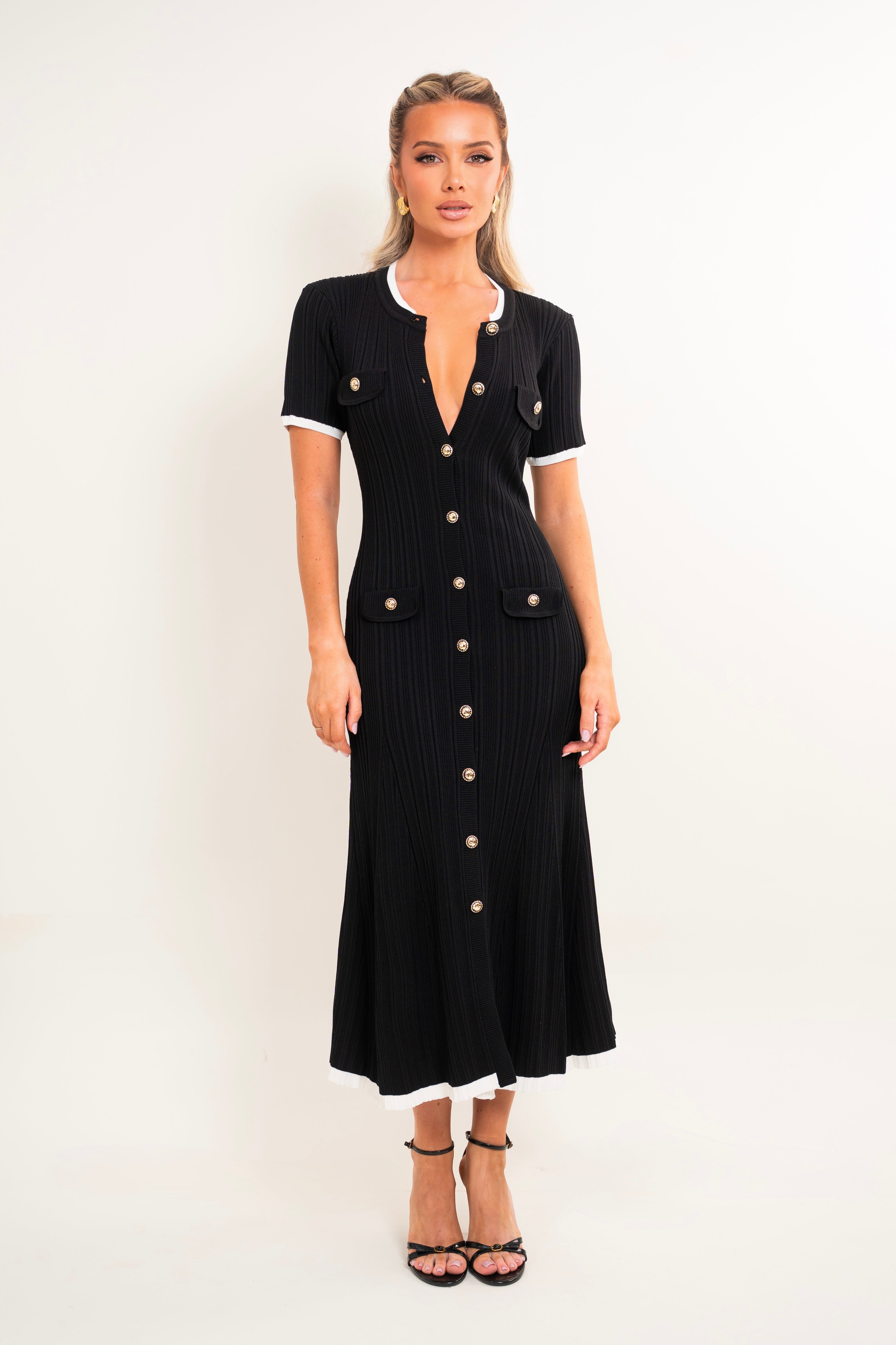 A woman modeling a black ribbed short-sleeve midi dress with white contrast trim at the collar, sleeve cuffs, and hem, featuring gold-tone buttons down the front and on the chest pockets, set against a plain white studio background.