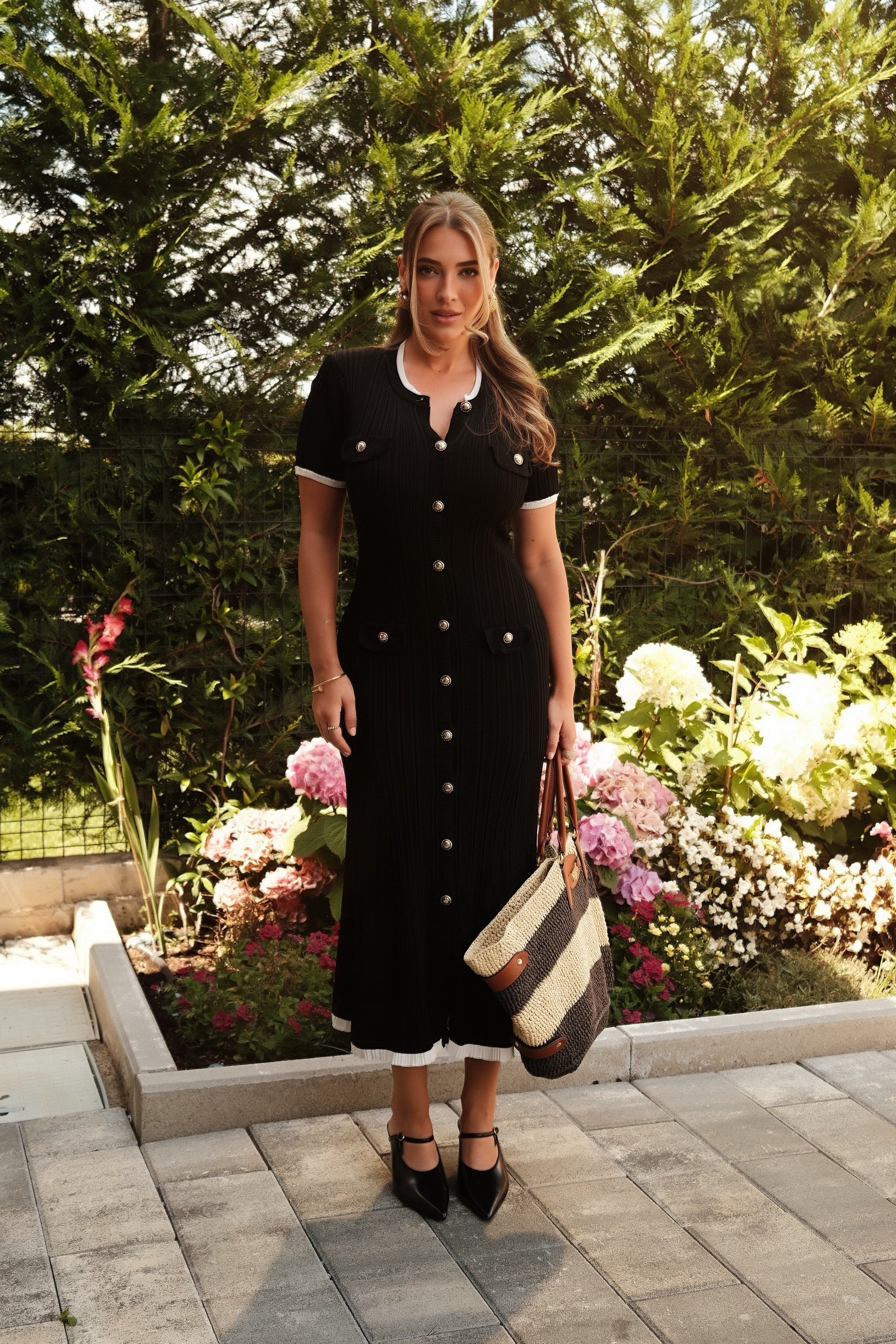 A woman stands outdoors in a garden setting wearing a black midi dress with white trim and button details, holding a striped tote bag.