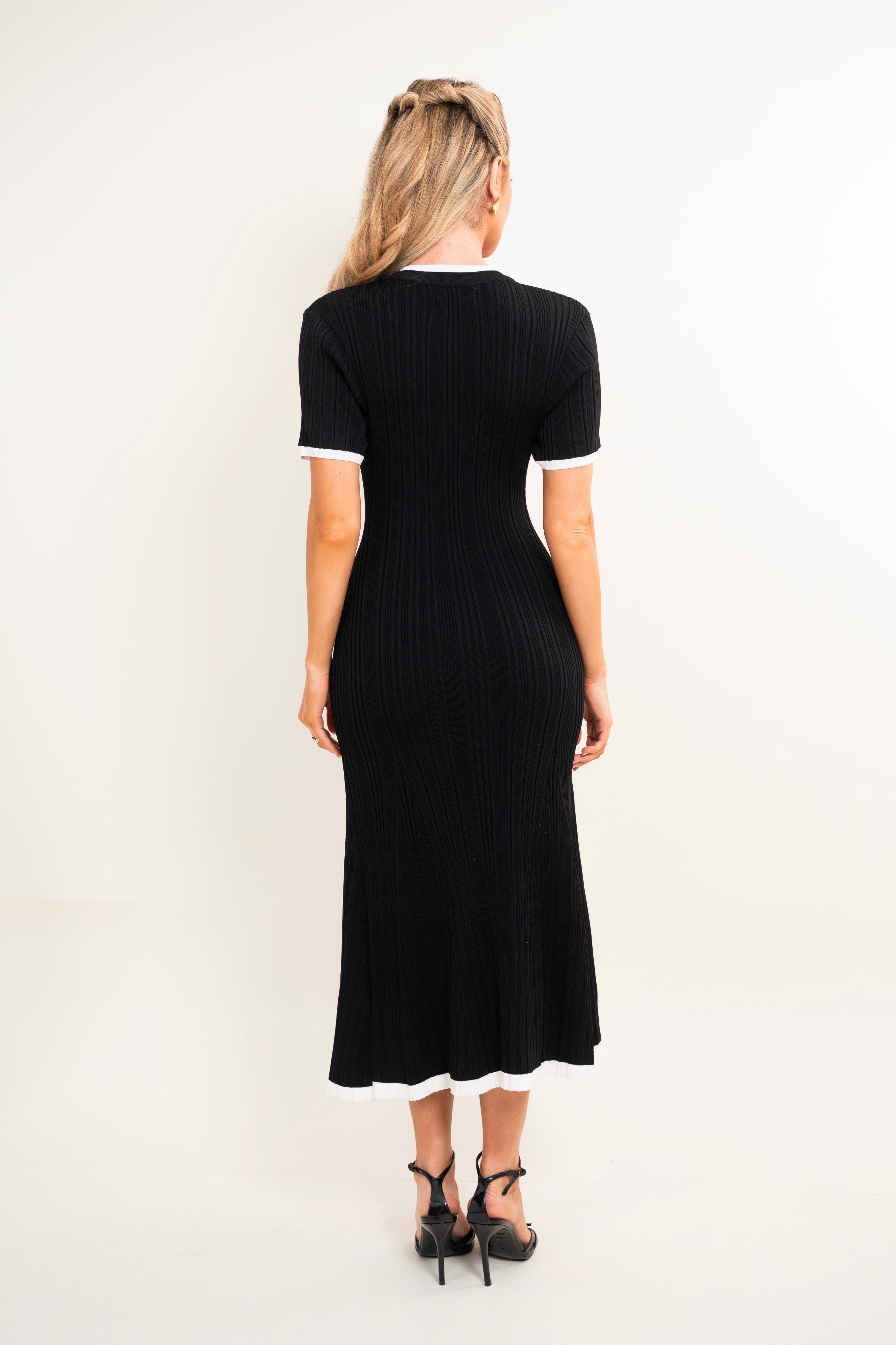A woman viewed from the back wearing a black ribbed short-sleeve midi dress with white trim at the collar and sleeve cuffs, paired with black high heels, against a plain white studio background.