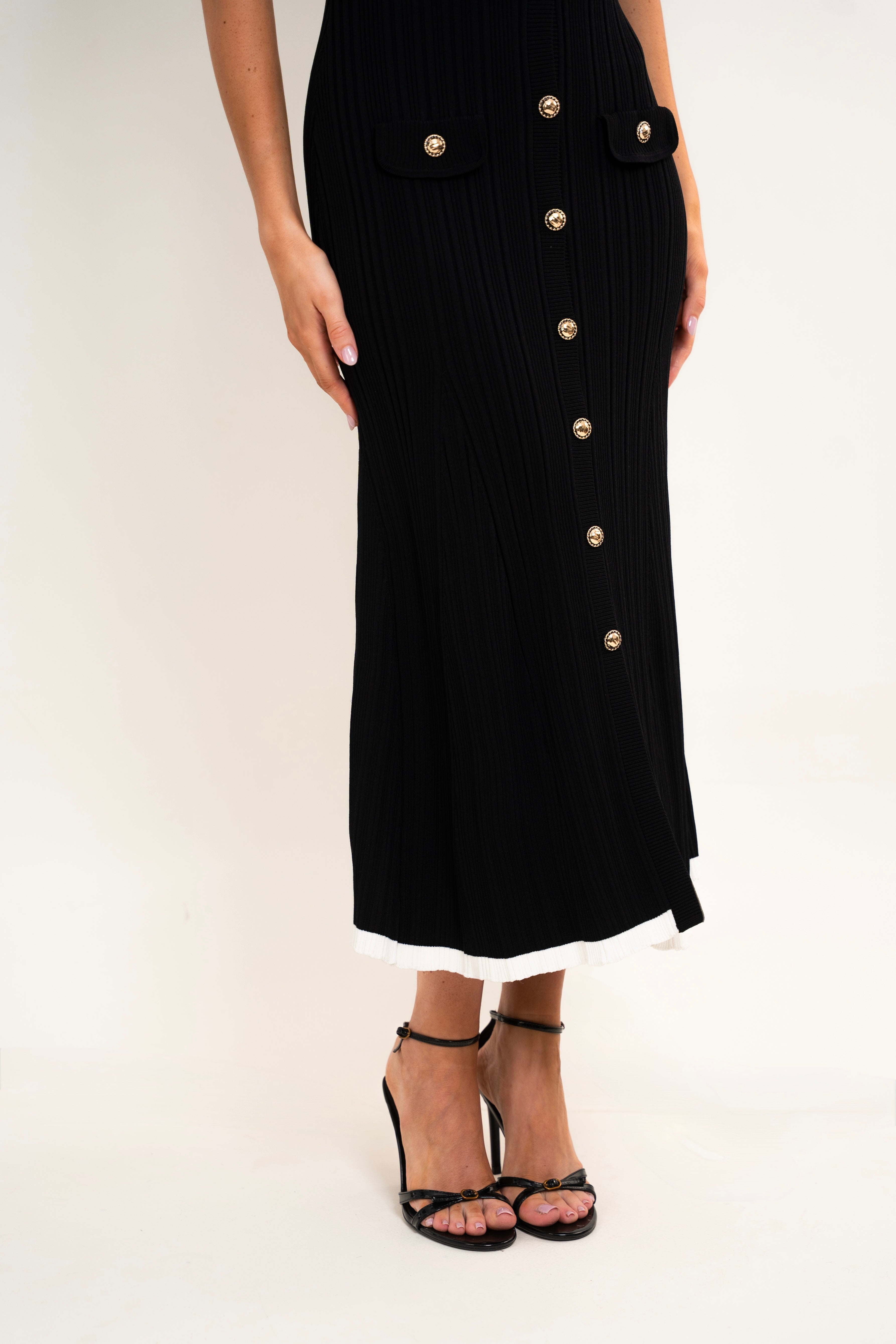 A model wearing a black ribbed knit midi skirt with gold-tone decorative buttons down the front and flap pockets, paired with black strappy high-heeled sandals against a plain light background.