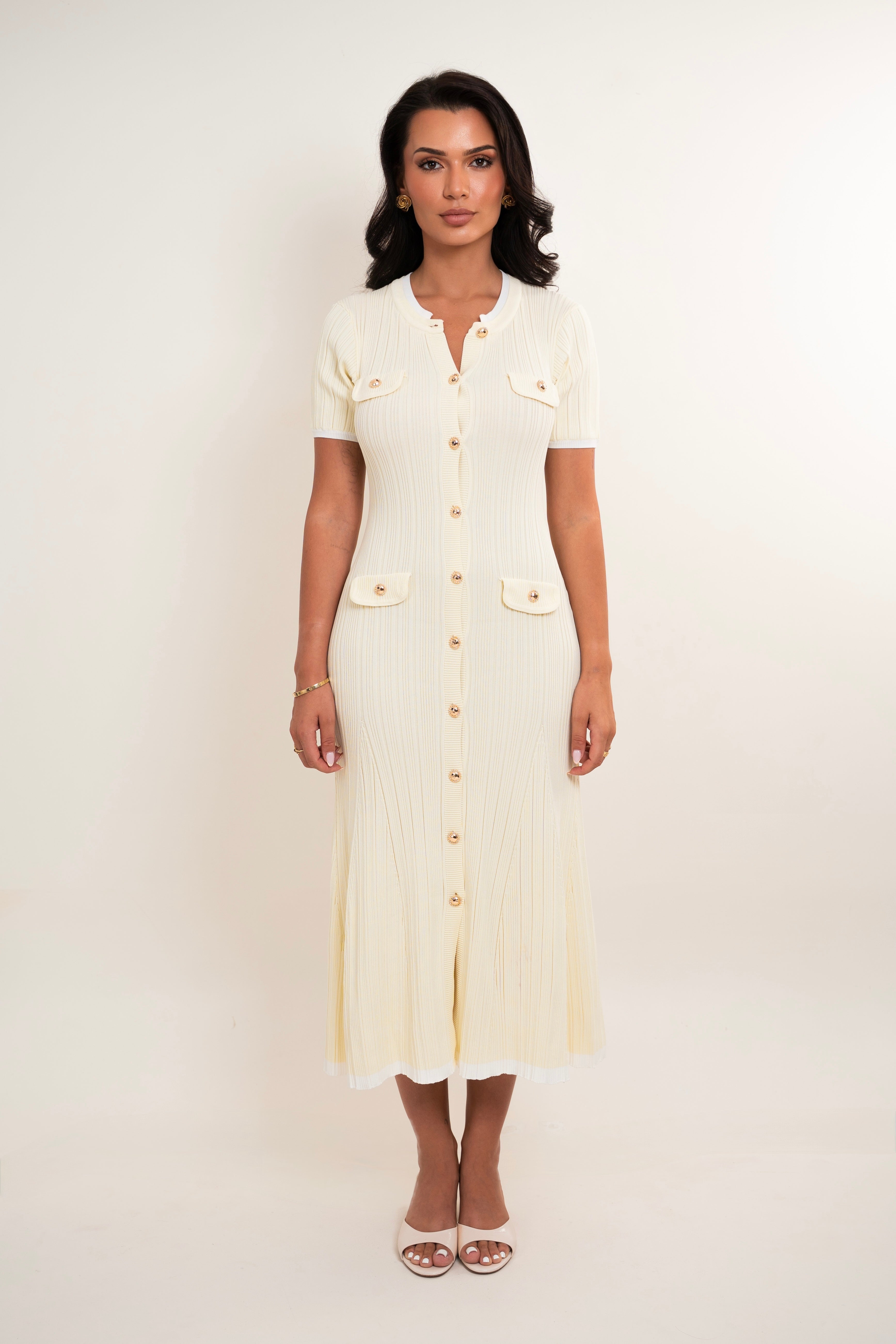 A woman modeling a cream-colored short-sleeve ribbed midi dress with gold buttons and flap pockets, standing against a plain off-white studio background.