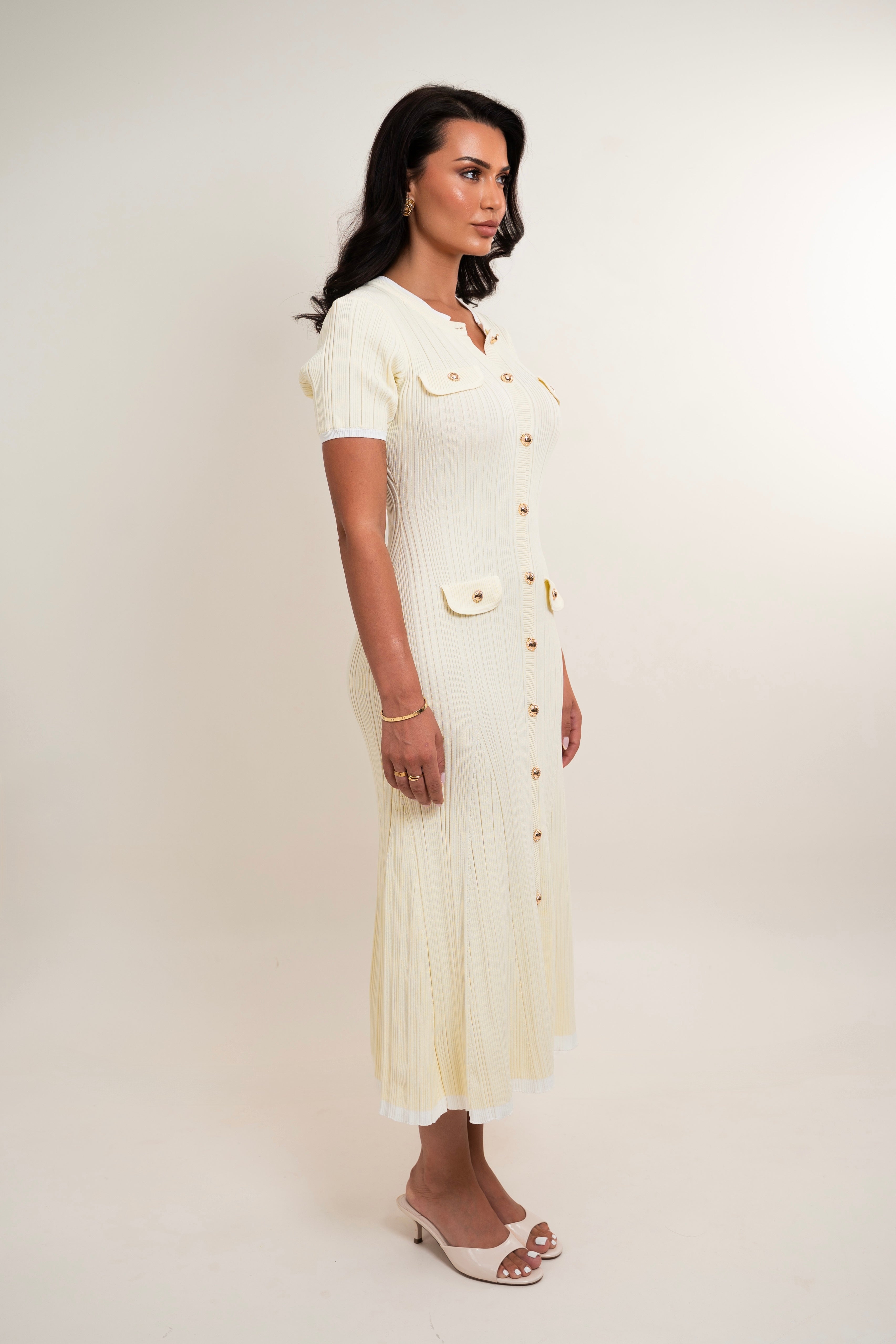 A woman modeling a cream-colored pleated midi dress with short sleeves, button details, and flap pockets, standing against a plain light background.