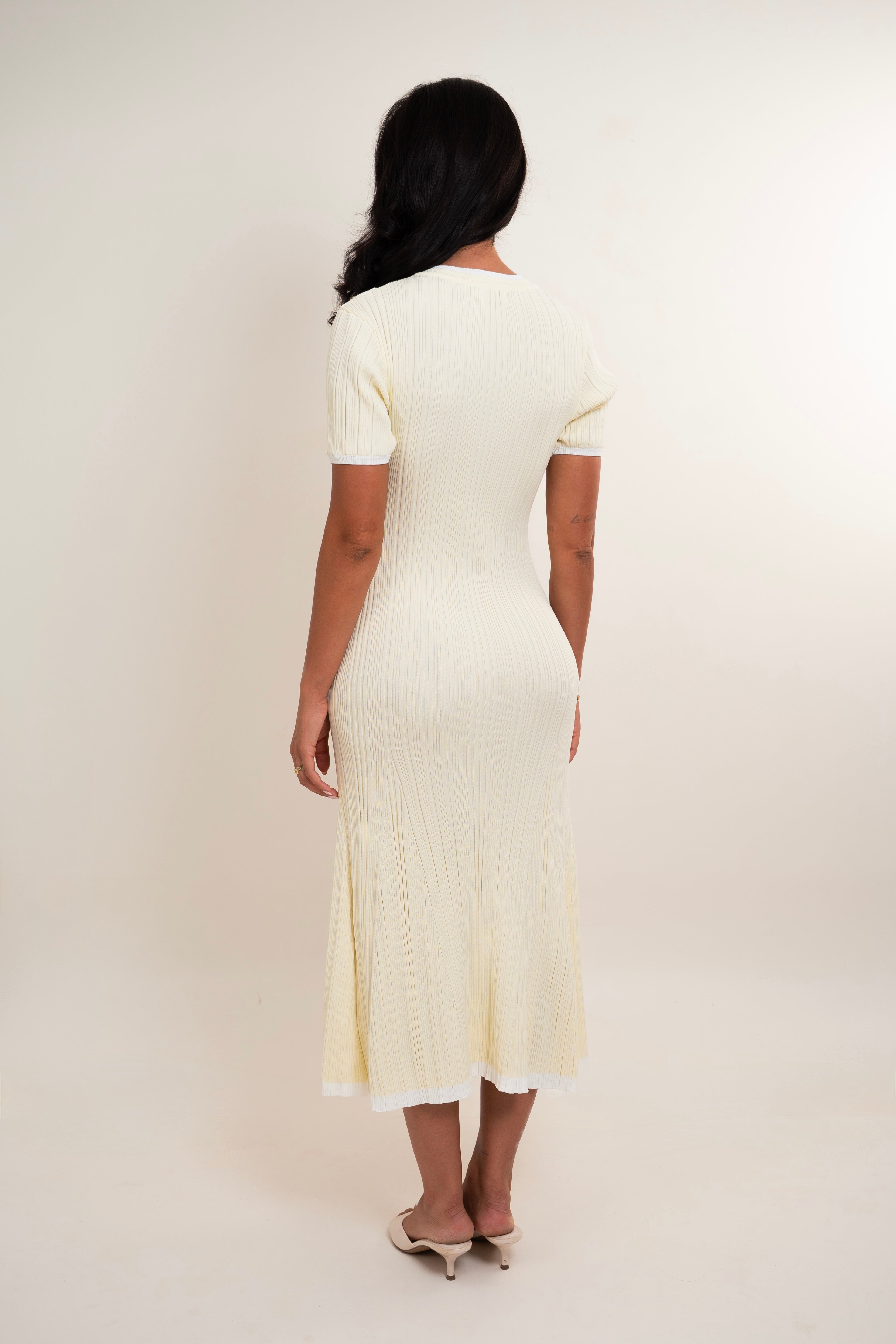 A woman viewed from the back wearing a short-sleeve cream ribbed midi dress and beige pointed-toe heels against a plain light beige studio background.