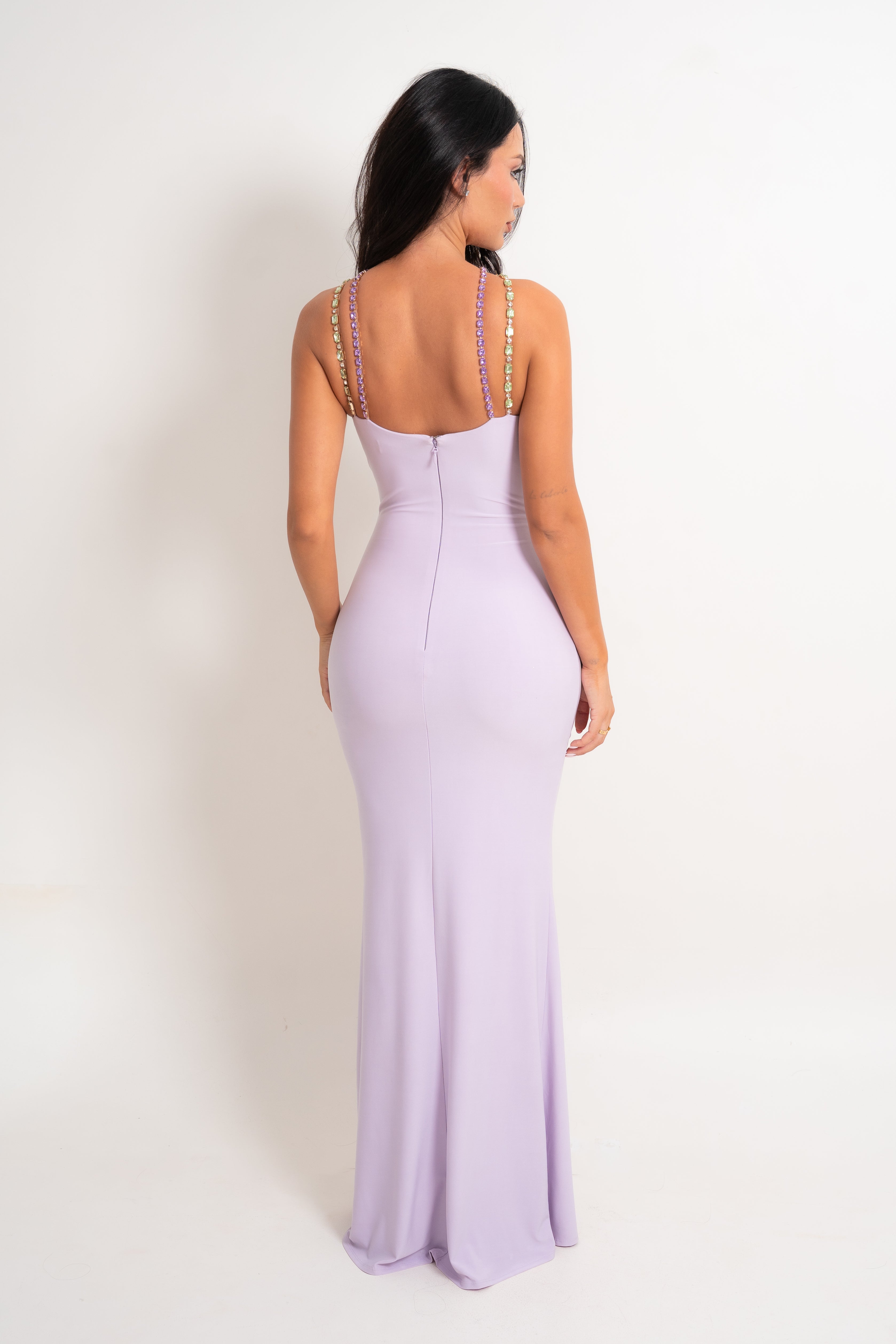 A woman shown from the back wearing a lavender floor-length fitted dress with thin embellished straps, photographed against a plain light background.