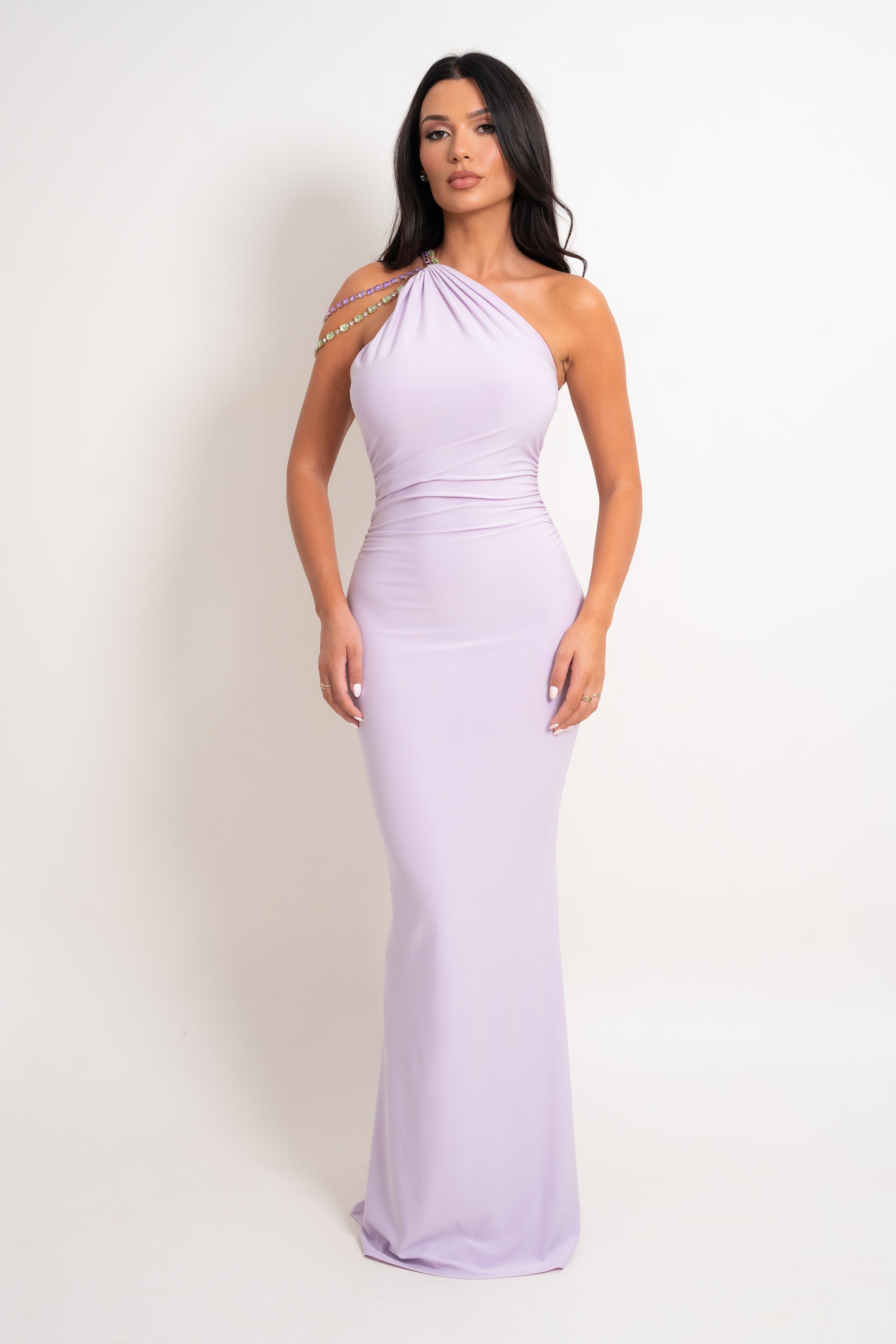 A woman modeling a lavender one-shoulder maxi dress with a crystal-embellished shoulder strap, standing against a plain white studio background.