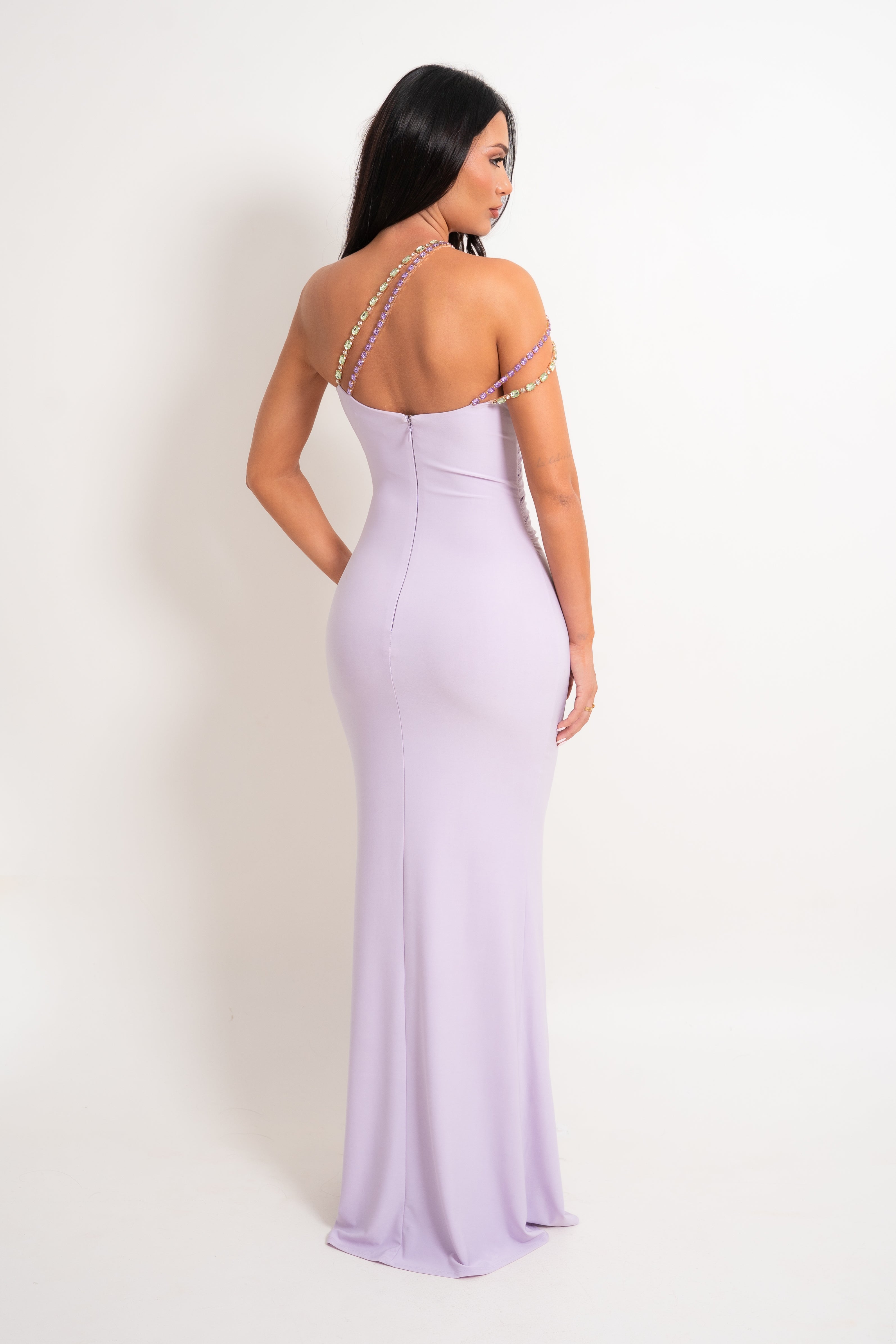 A woman modeling a lavender off-the-shoulder maxi dress from the back, featuring thin rhinestone-embellished straps and a fitted silhouette, set against a plain white studio background.