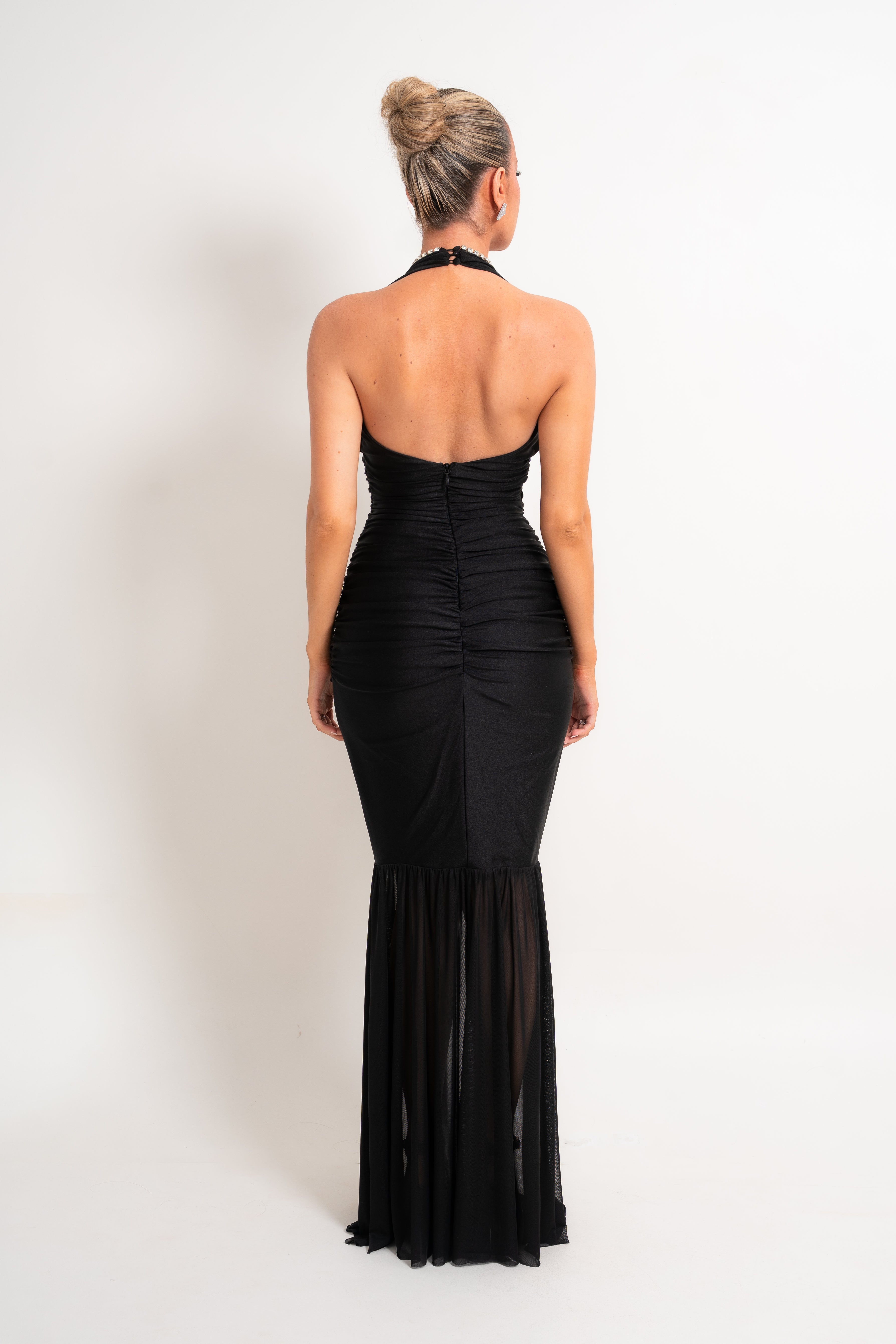 A woman viewed from the back wearing a black halter-neck, ruched mermaid gown against a plain white studio background.