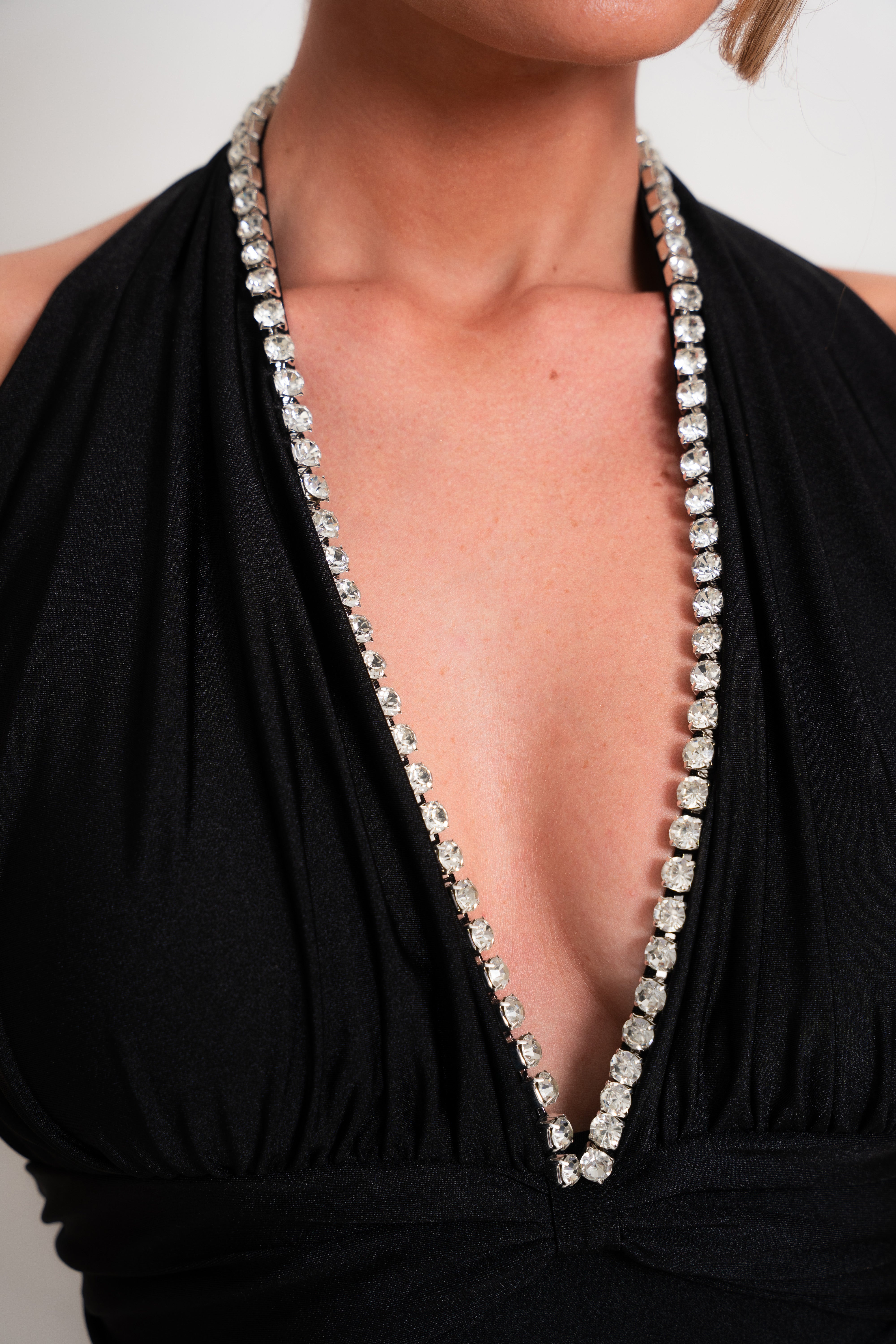 A close-up of a model wearing a black halter-neck dress with a very deep V-neckline, embellished with a long, sparkling silver rhinestone border along the neckline.