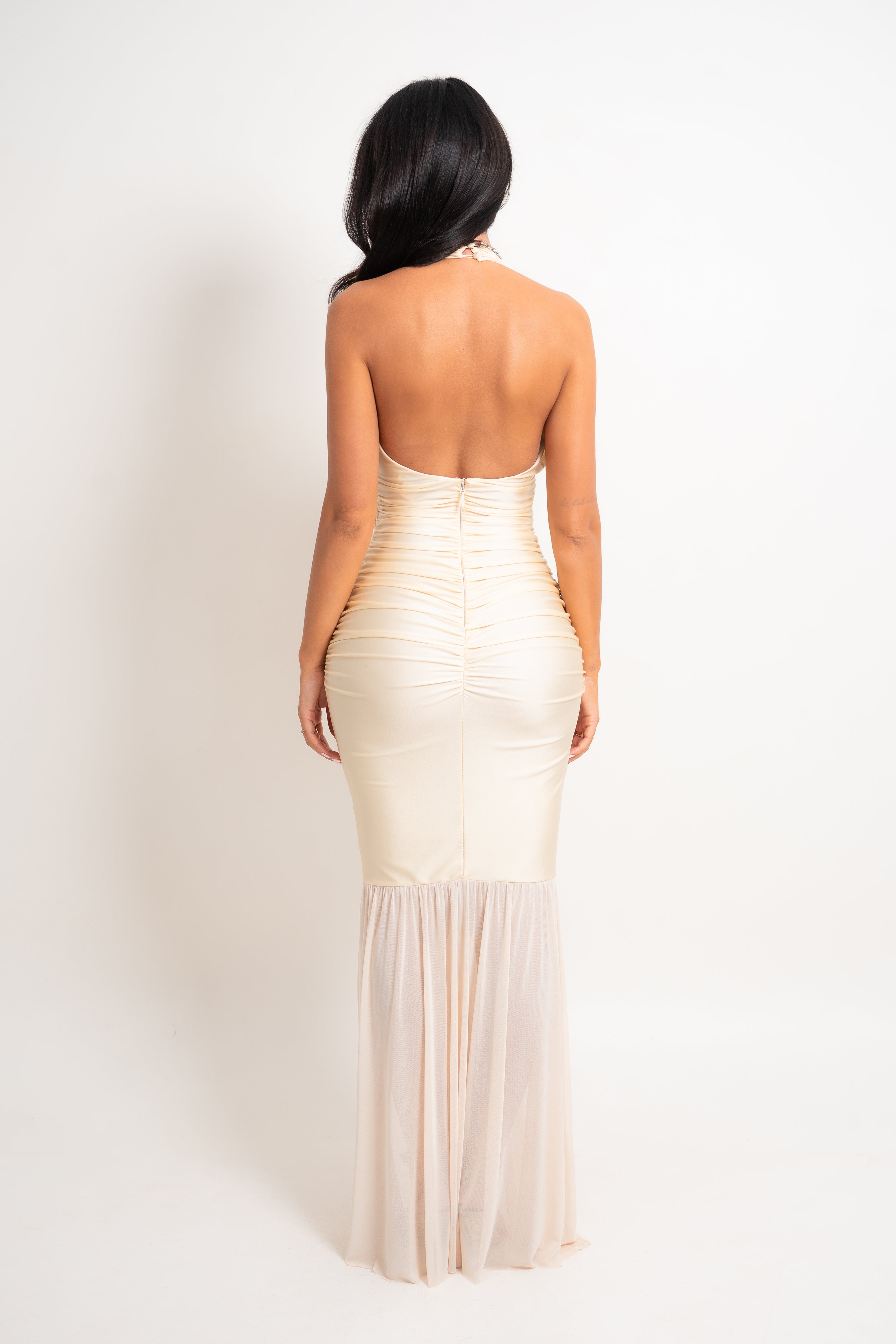A woman viewed from the back wearing a champagne-colored, strapless ruched mermaid gown with a sheer mesh lower skirt, set against a plain white studio background.