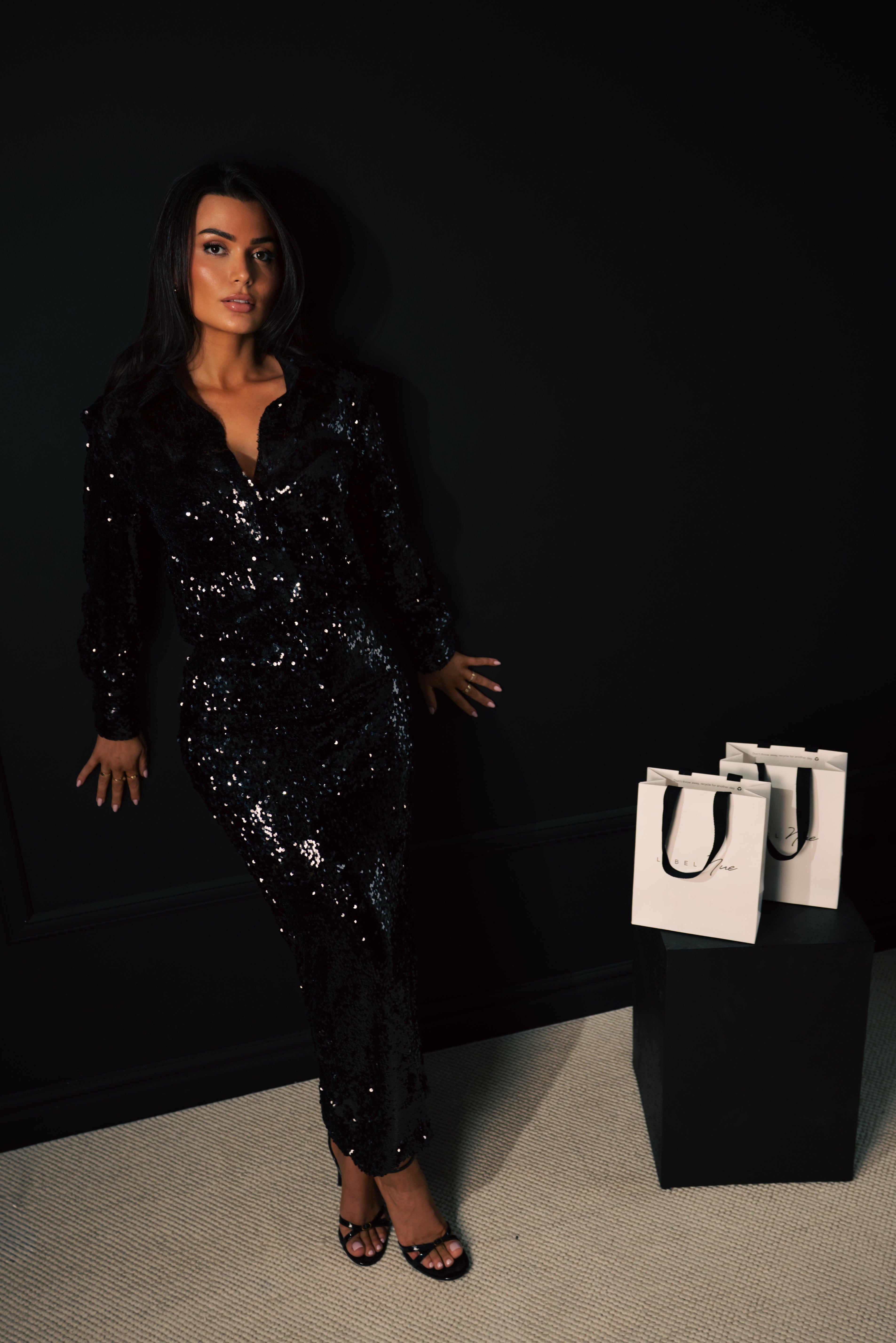 Woman in a black sequin outfit standing next to white bags against a dark background