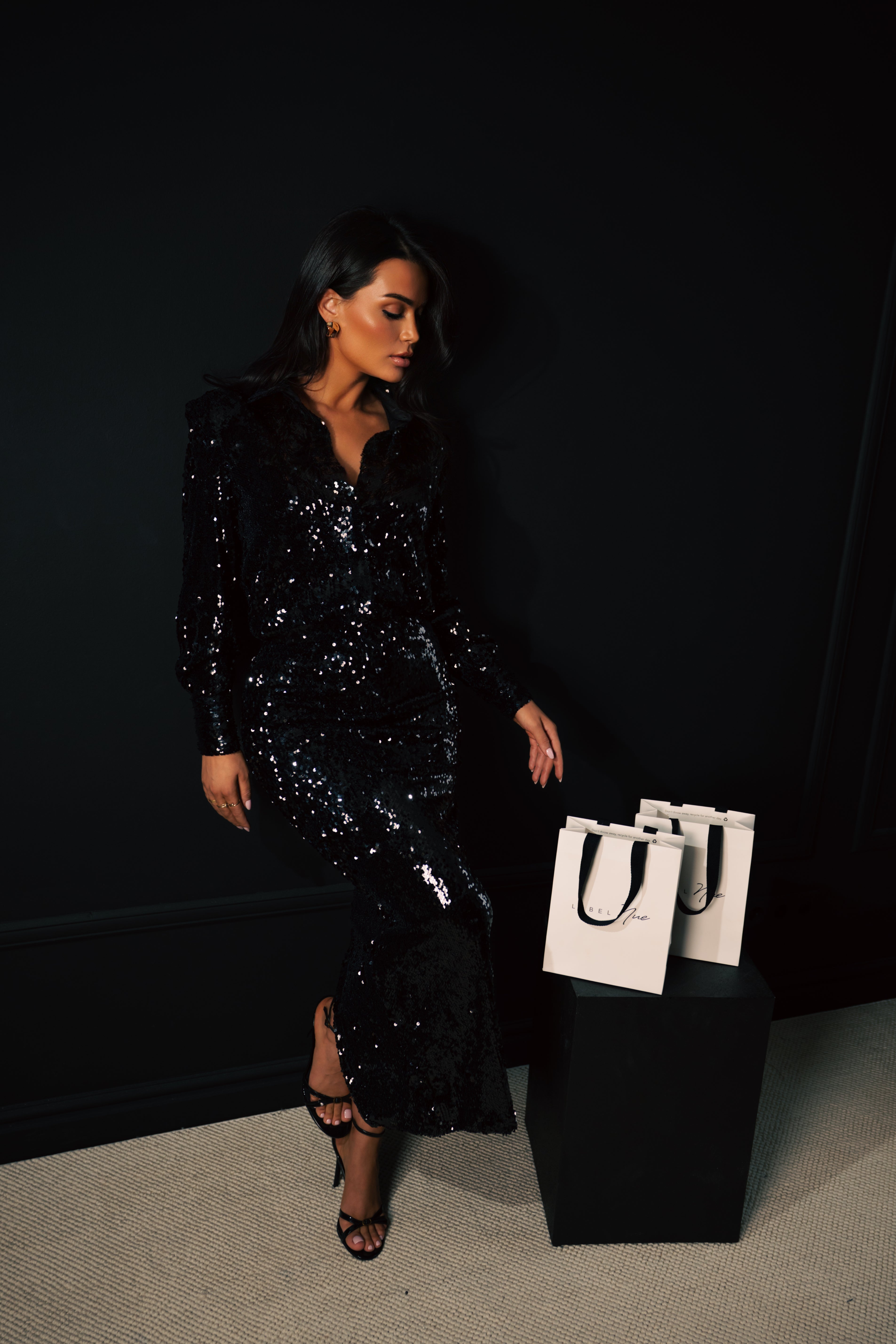 Woman in a black sequin dress standing next to two white handbags against a dark background