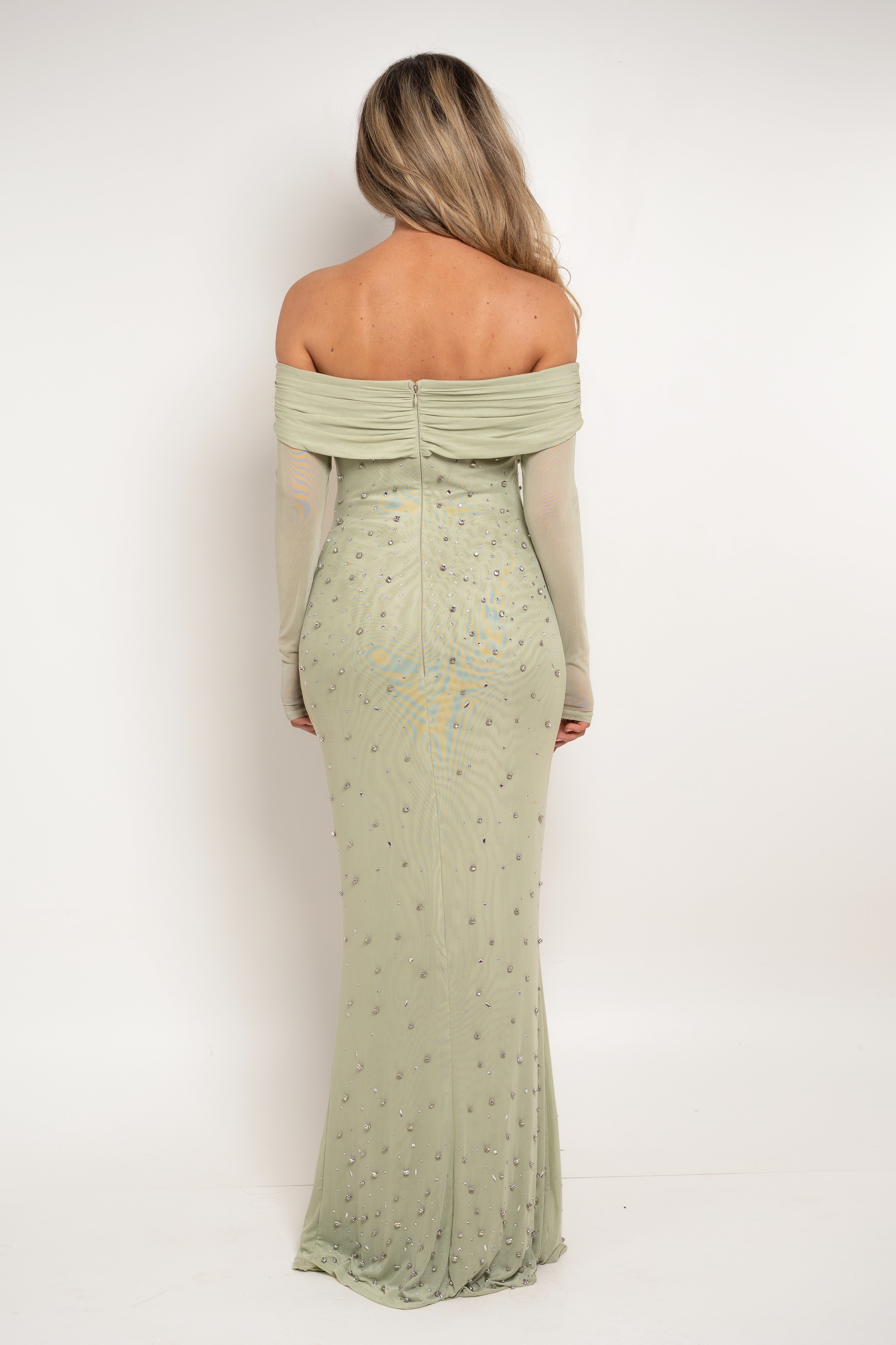 A woman viewed from the back wearing a sage green off-the-shoulder maxi dress with sheer long sleeves and scattered rhinestone embellishments, set against a plain white studio background.