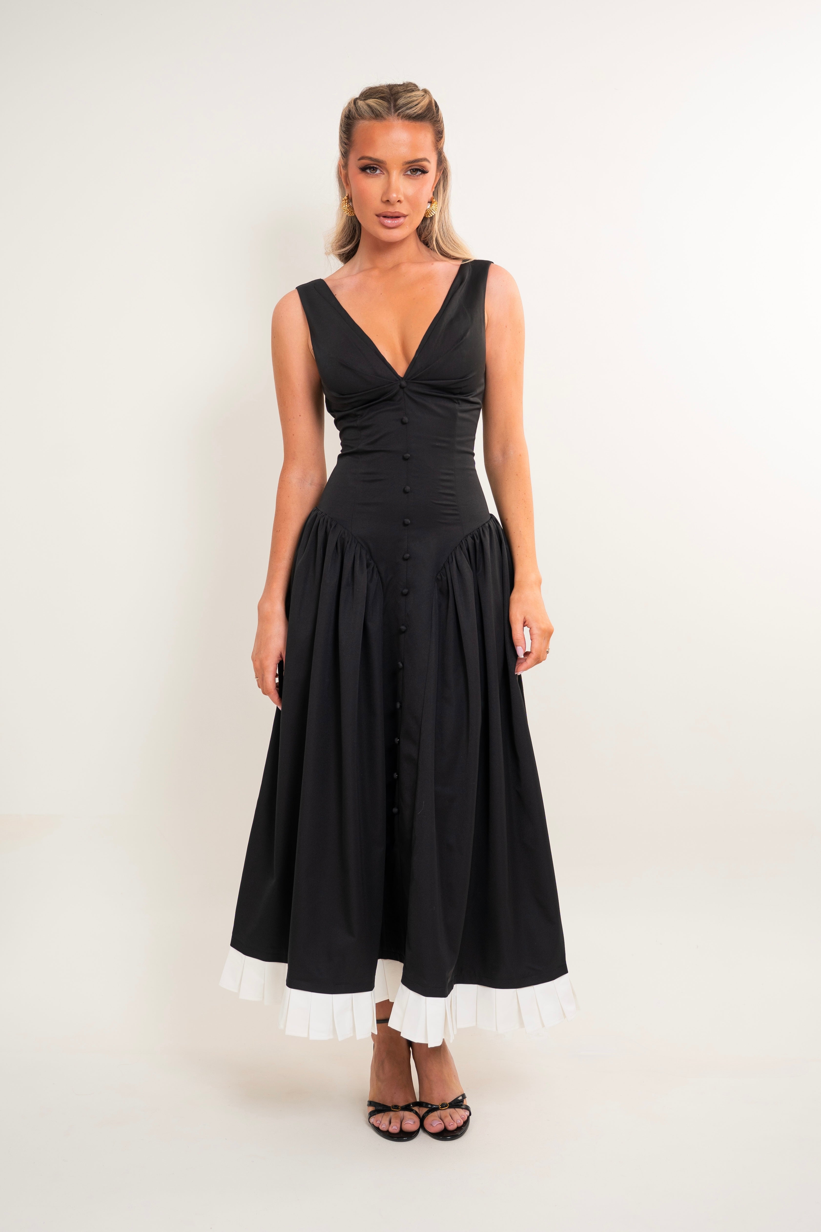 A woman modeling a sleeveless black midi dress with a deep V-neckline, buttoned bodice, and a white ruffled hem trim, standing against a plain off-white studio background.