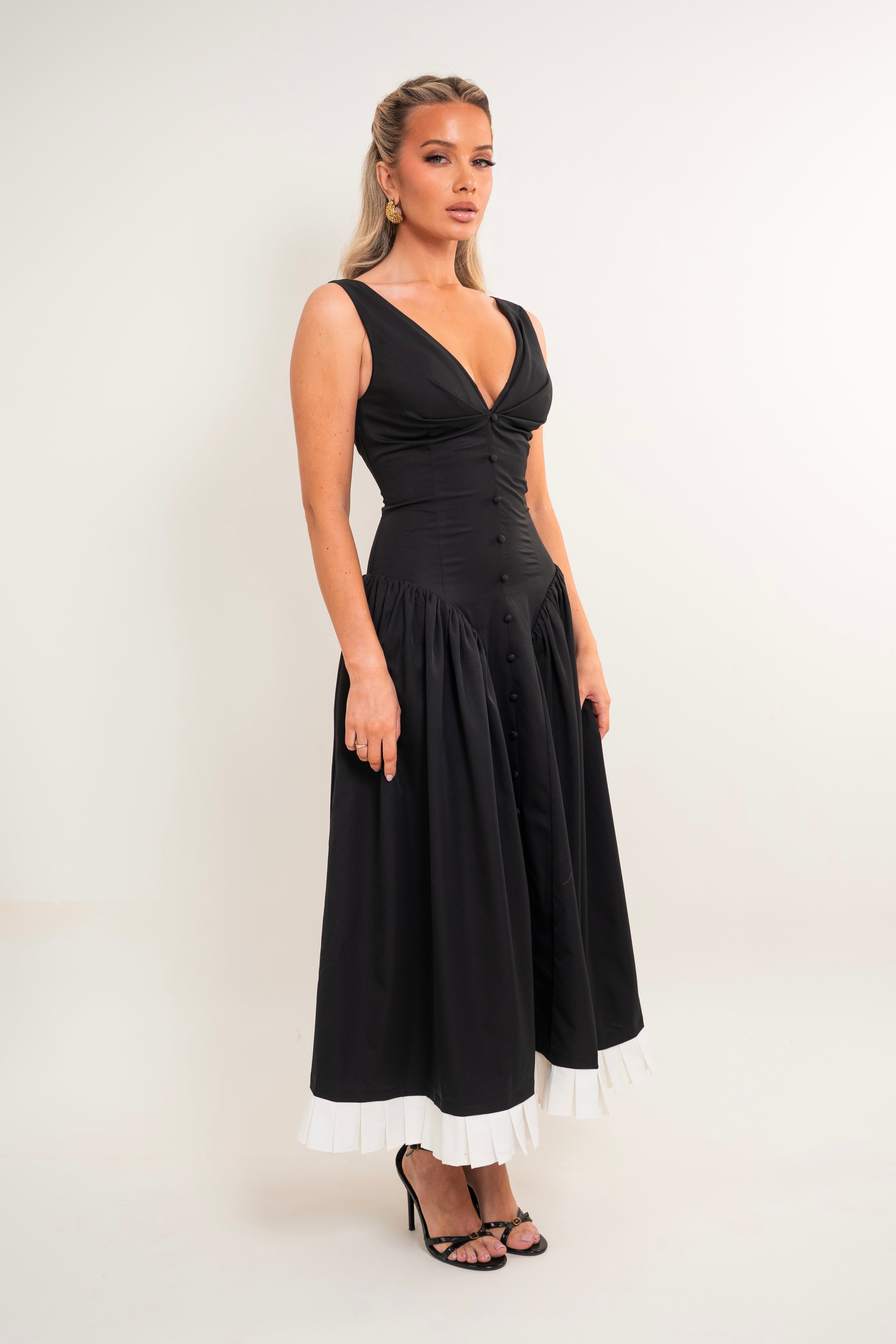 A woman modeling a sleeveless black midi dress with a deep V-neckline, gathered waist, buttoned bodice, and a contrasting white ruffled hem, set against a plain light background.