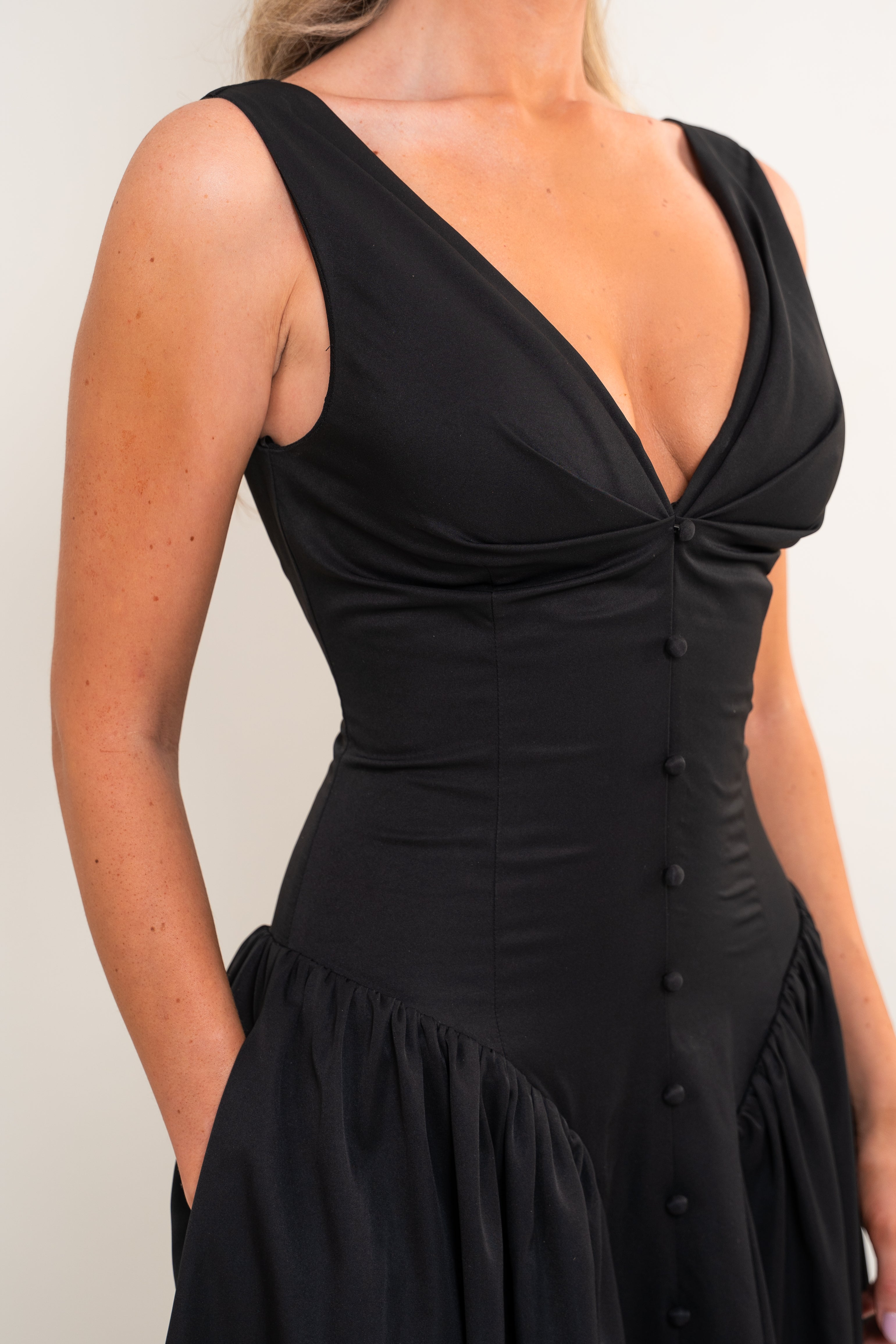 A close-up of a woman wearing a black sleeveless dress with a deep V-neckline, gathered bust, button front, and flared skirt against a plain light background.