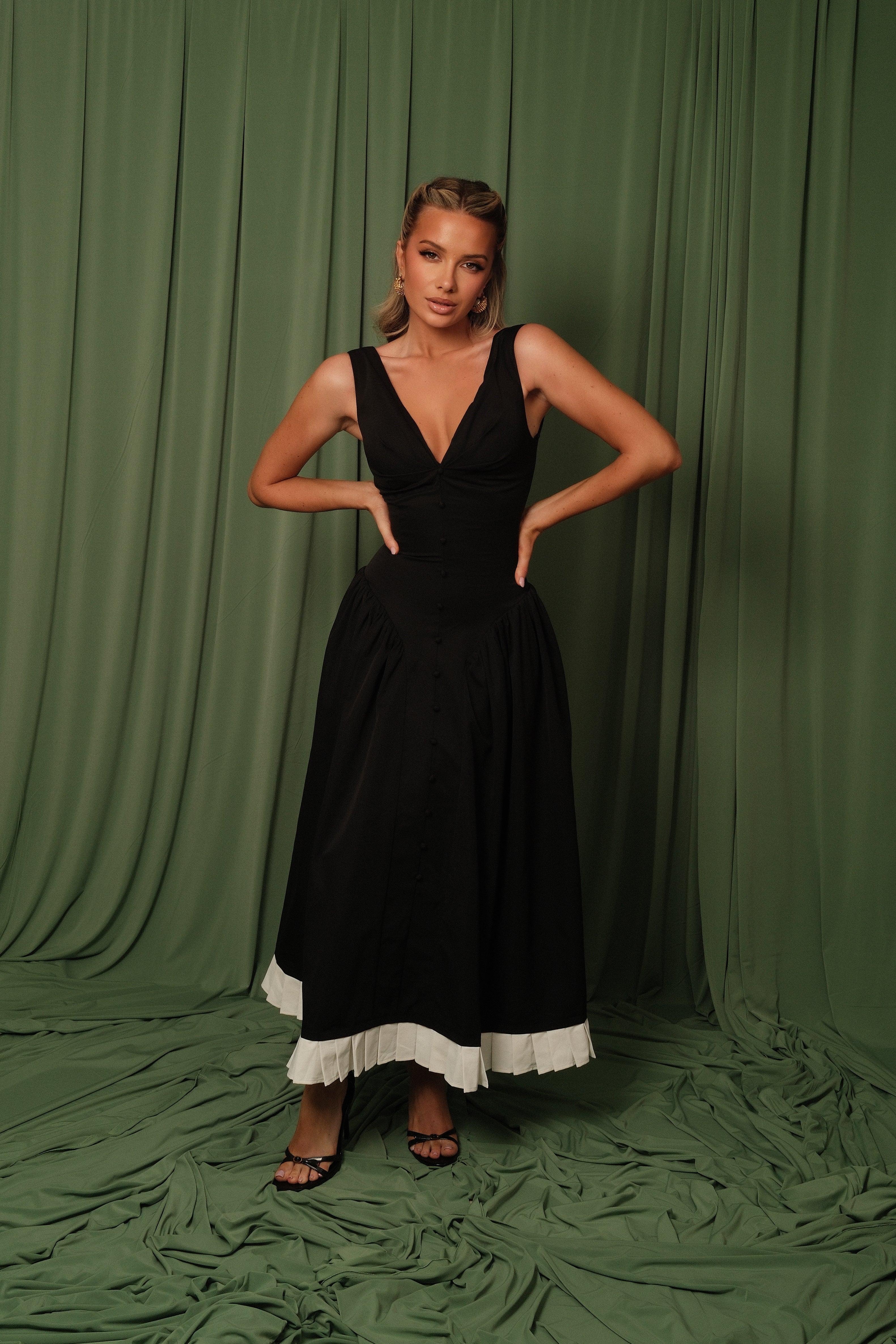 A woman modeling a black sleeveless maxi dress with a white ruffled hem, standing against a draped olive green fabric backdrop.