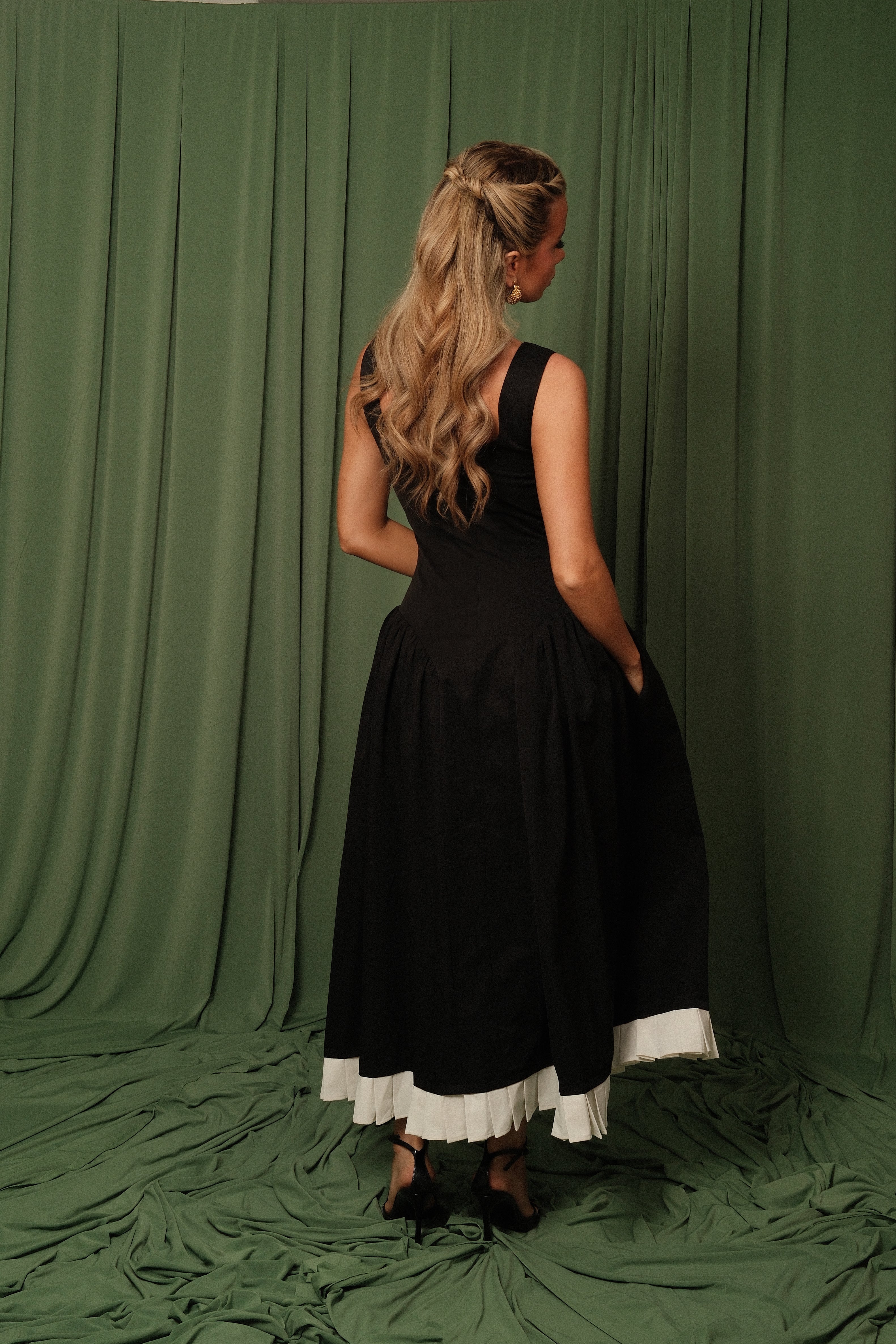 A woman stands with her back to the camera wearing a sleeveless black midi dress with a contrasting white ruffled hem, posed in front of a draped olive green fabric backdrop.