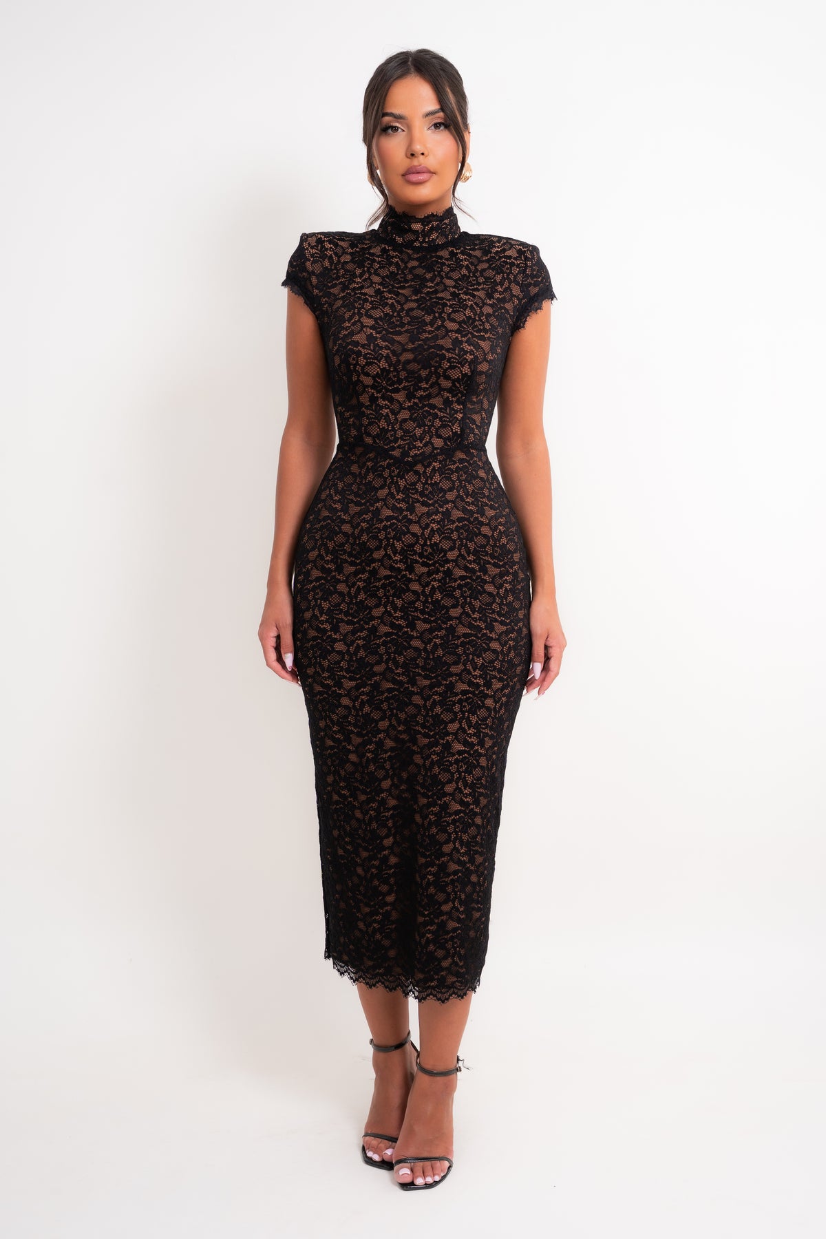 A woman modeling a black lace midi dress with cap sleeves and a high mock neck, standing against a plain white studio background.