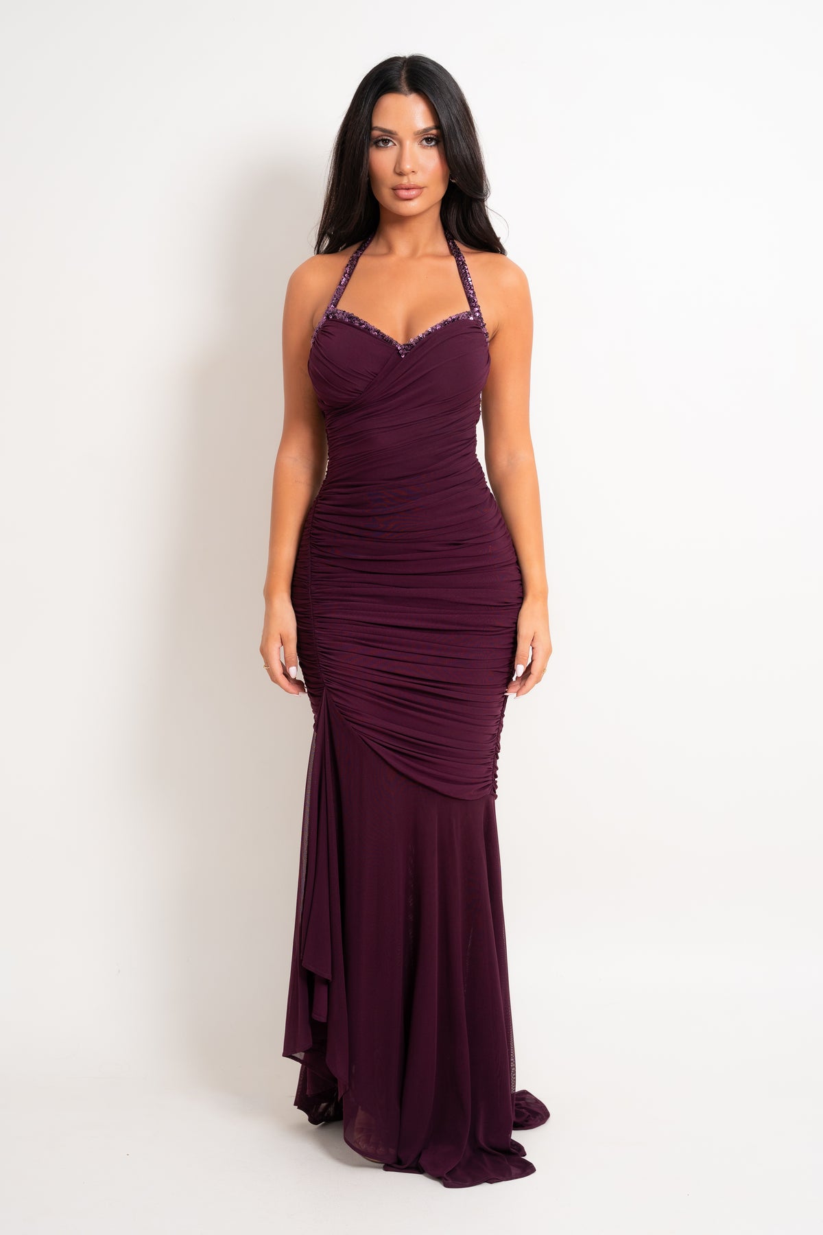 A woman modeling a floor-length halter gown in deep burgundy with ruched draping and a draped side detail, photographed against a plain light gray studio background.