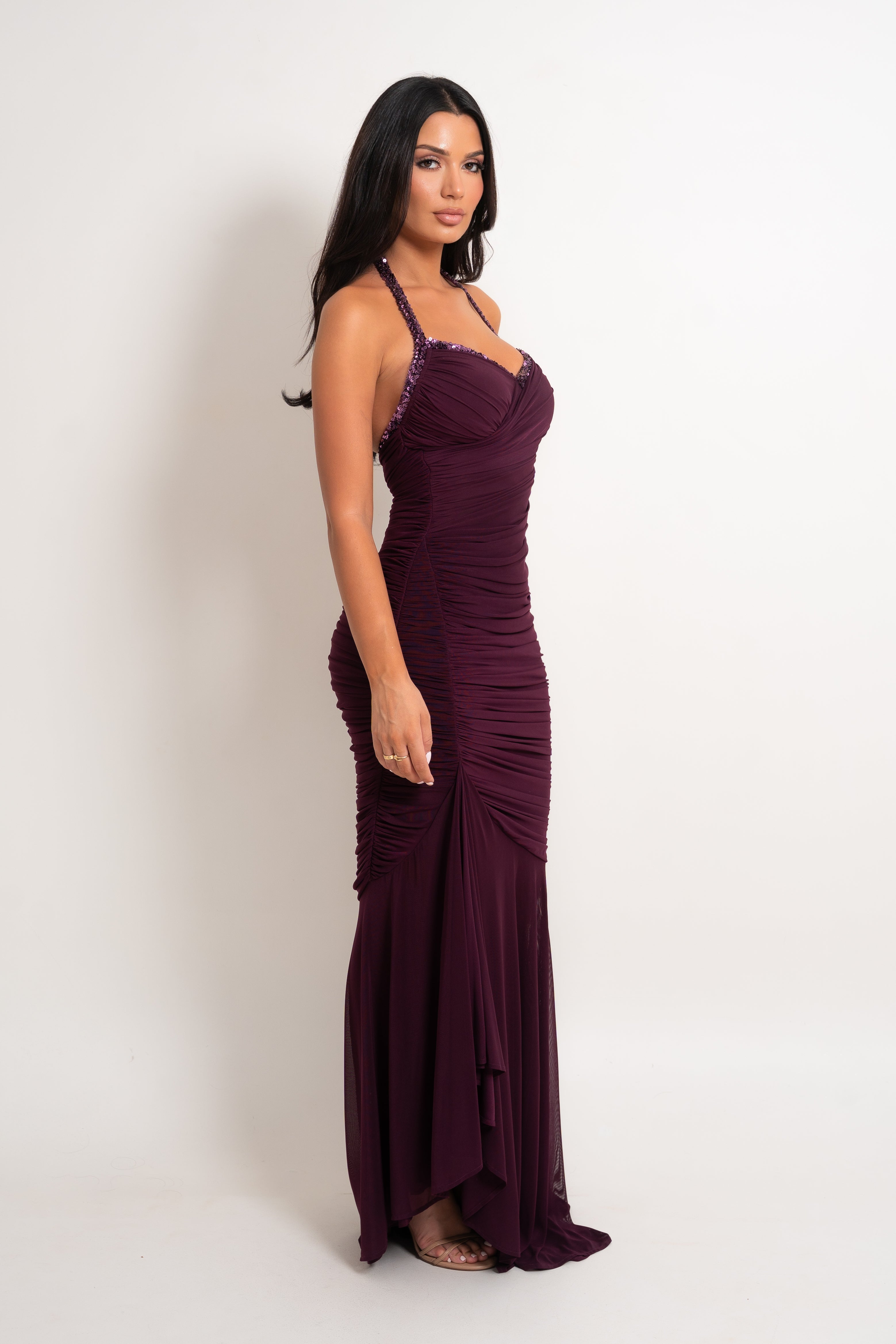 A woman modeling a floor-length, ruched evening gown in deep burgundy with thin shoulder straps and a sweetheart neckline, photographed against a plain light studio background.